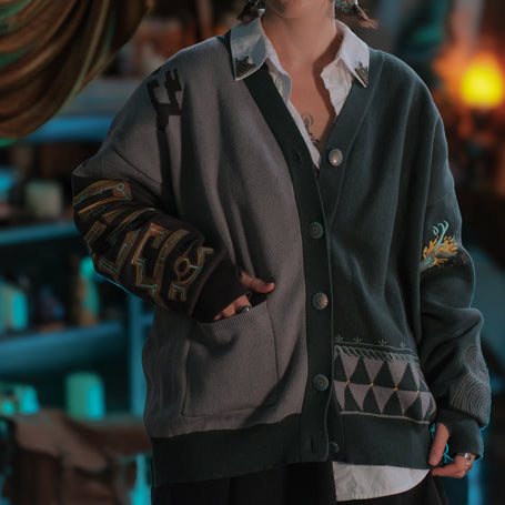 The Archaic Cardigan is an oversized 100% cotton knit cardigan in dark turquoise, slate grey, and tarnished gold. It features two front pockets and embroidered details througout.