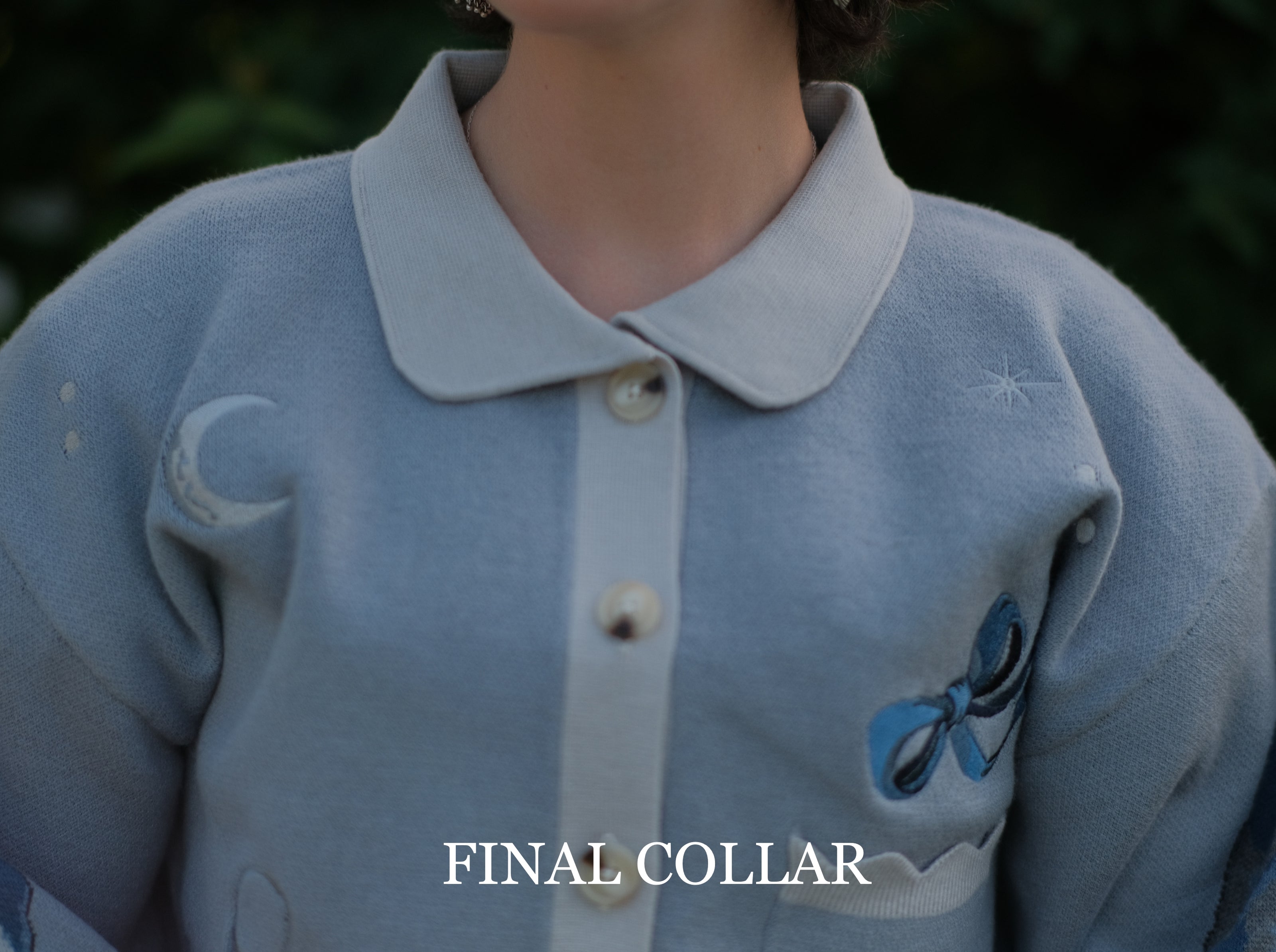 A soft, knit, cropped cardigan, in tones of dove-grey and dusty-blue. This cardigan features detailed embroidery of a bow, moon and stars, stork scissors, and bluebird wings on both sleeves. The cardigan also features red-ribbon drawstring sleeves for customization, and scalloped edge detailing.