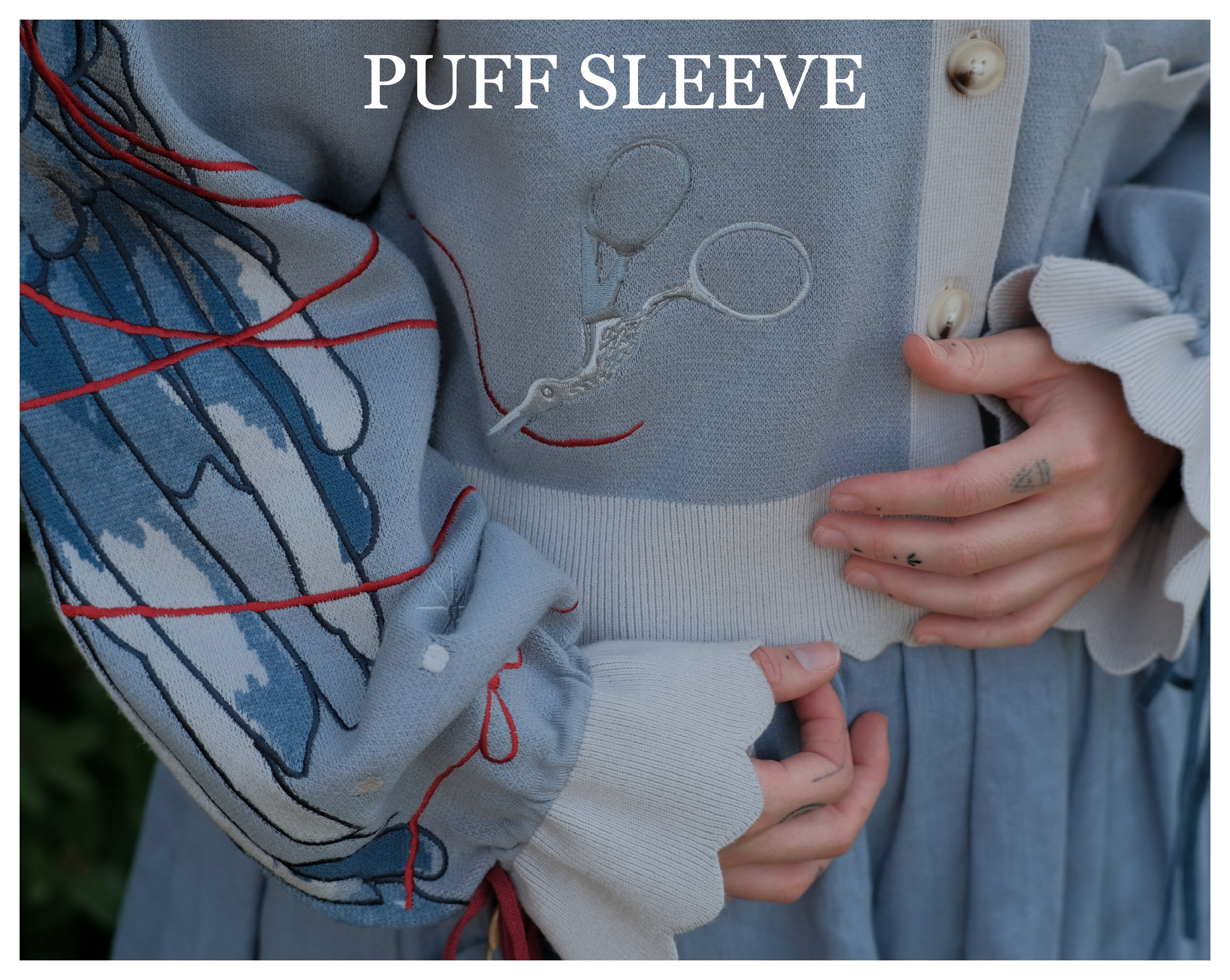 A soft, knit, cropped cardigan, in tones of dove-grey and dusty-blue. This cardigan features detailed embroidery of a bow, moon and stars, stork scissors, and bluebird wings on both sleeves. The cardigan also features red-ribbon drawstring sleeves for customization, and scalloped edge detailing.