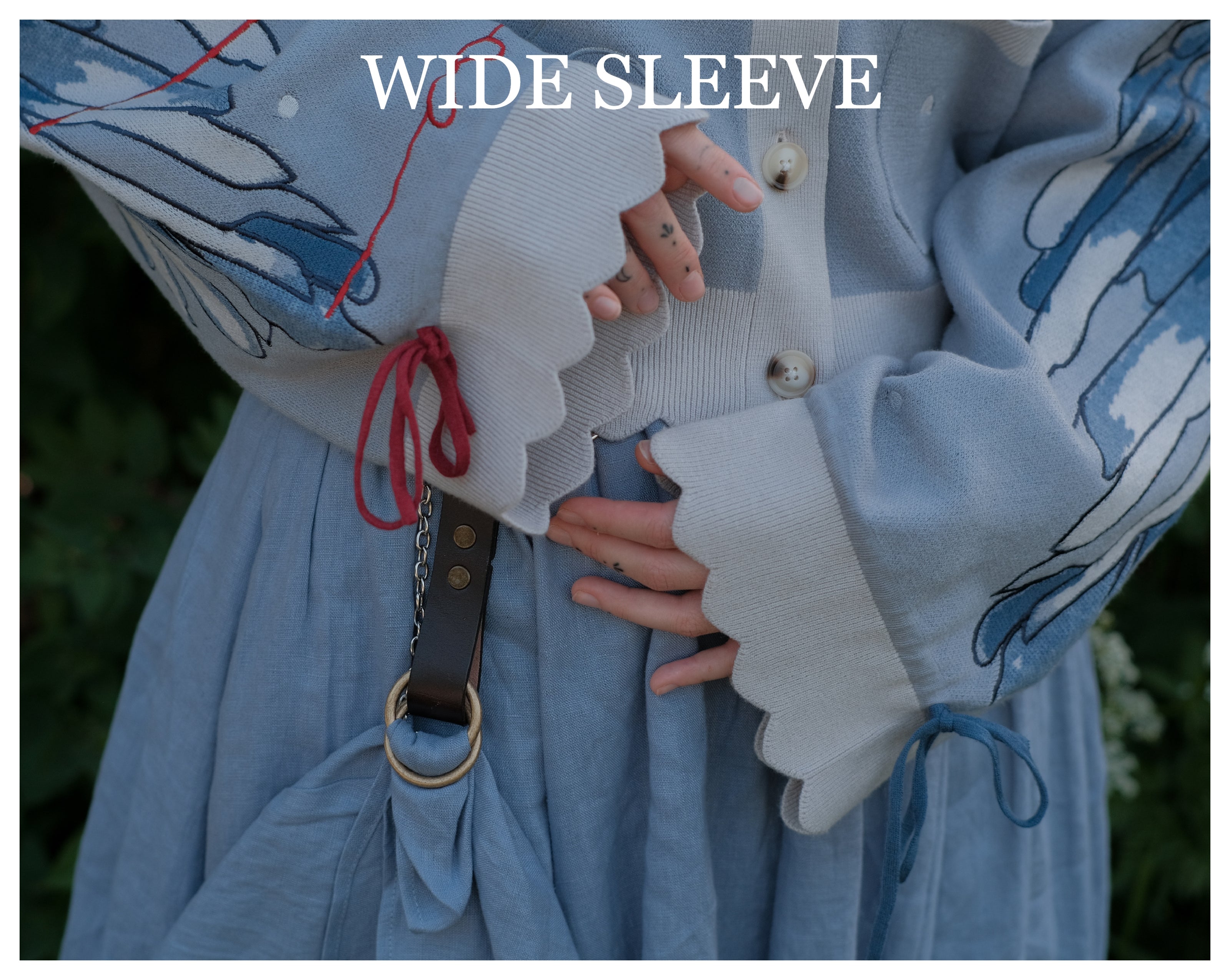 A soft, knit, cropped cardigan, in tones of dove-grey and dusty-blue. This cardigan features detailed embroidery of a bow, moon and stars, stork scissors, and bluebird wings on both sleeves. The cardigan also features red-ribbon drawstring sleeves for customization, and scalloped edge detailing.