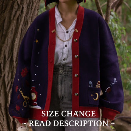 A soft, button-up, oversized, knit cardigan, in shades of navy blue, crimson red, and vintage gold. The cardigan features whimsical embroidered details of autumn leaves, moon and stars, a snowy lantern, and a bluebirds feather.