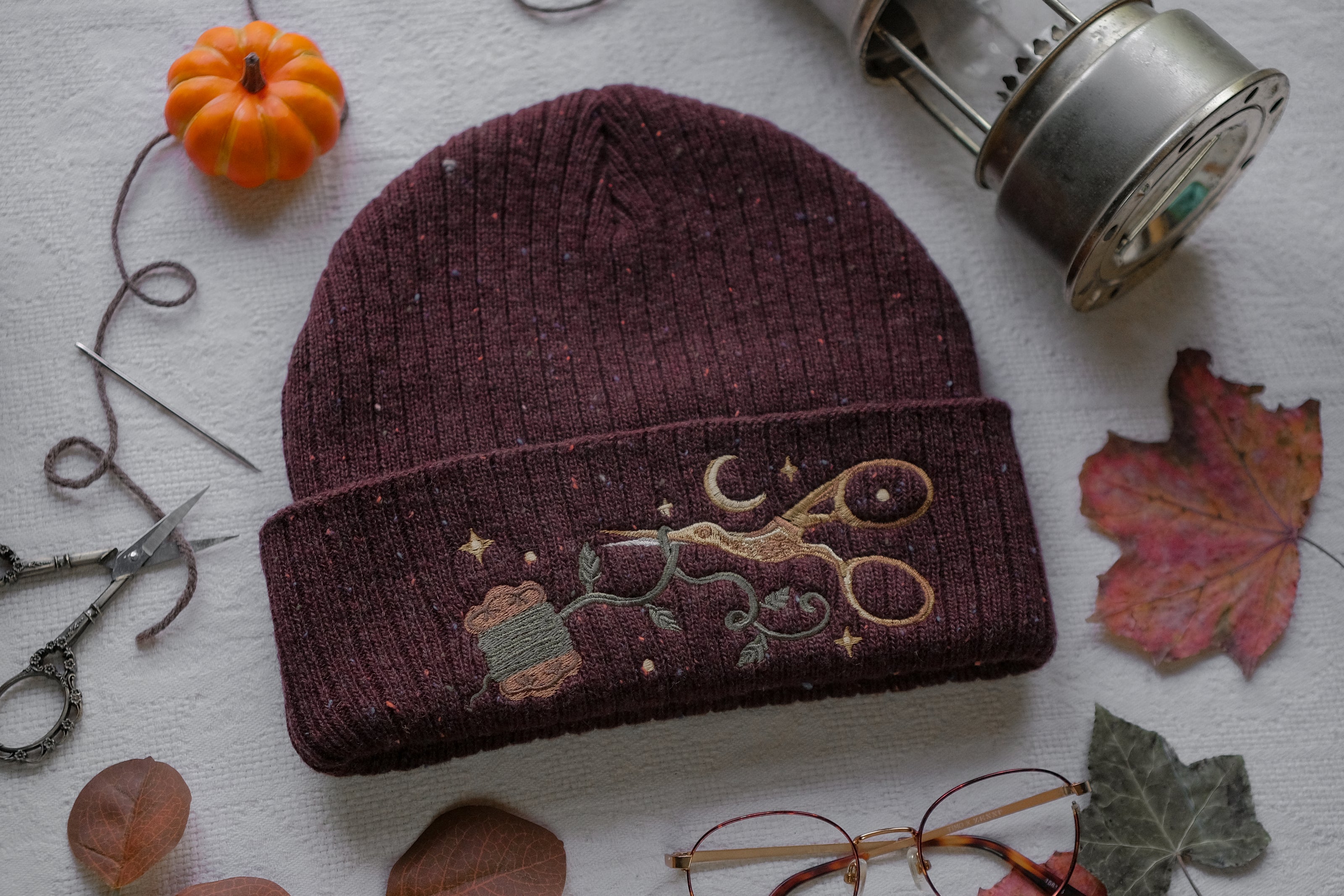 A cozy, flecked, amaranth-red, wool blend knit beanie, embroidered with a pair of golden, stork-shaped sewing scissors, and looping bobbin of green thread, under a crescent moon.