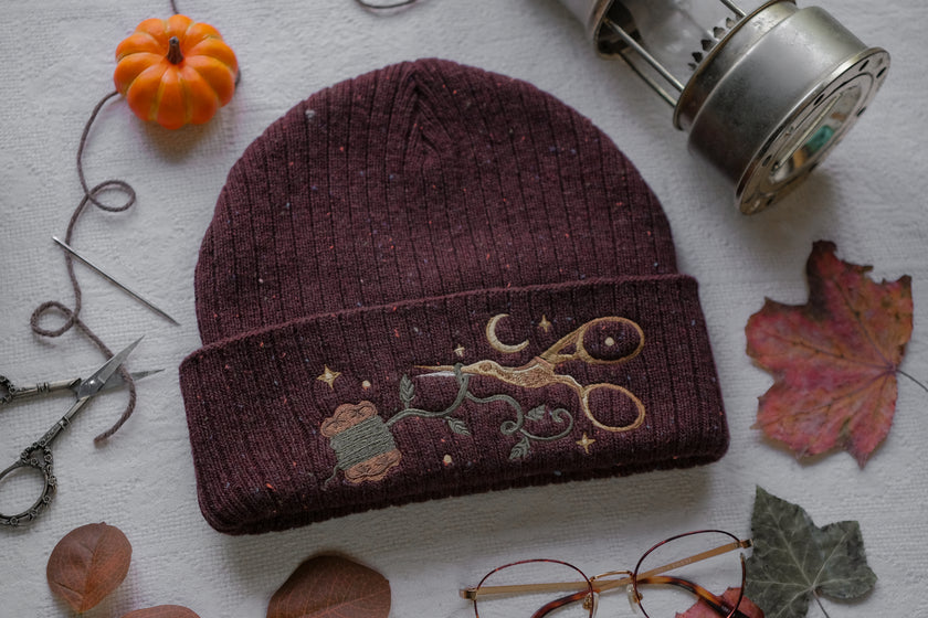 A cozy, flecked, amaranth-red, wool blend knit beanie, embroidered with a pair of golden, stork-shaped sewing scissors, and looping bobbin of green thread, under a crescent moon.