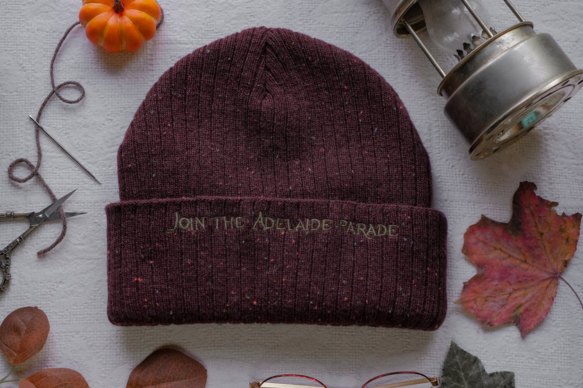 A cozy, flecked, amaranth-red, wool blend knit beanie, embroidered with a pair of golden, stork-shaped sewing scissors, and looping bobbin of green thread, under a crescent moon.