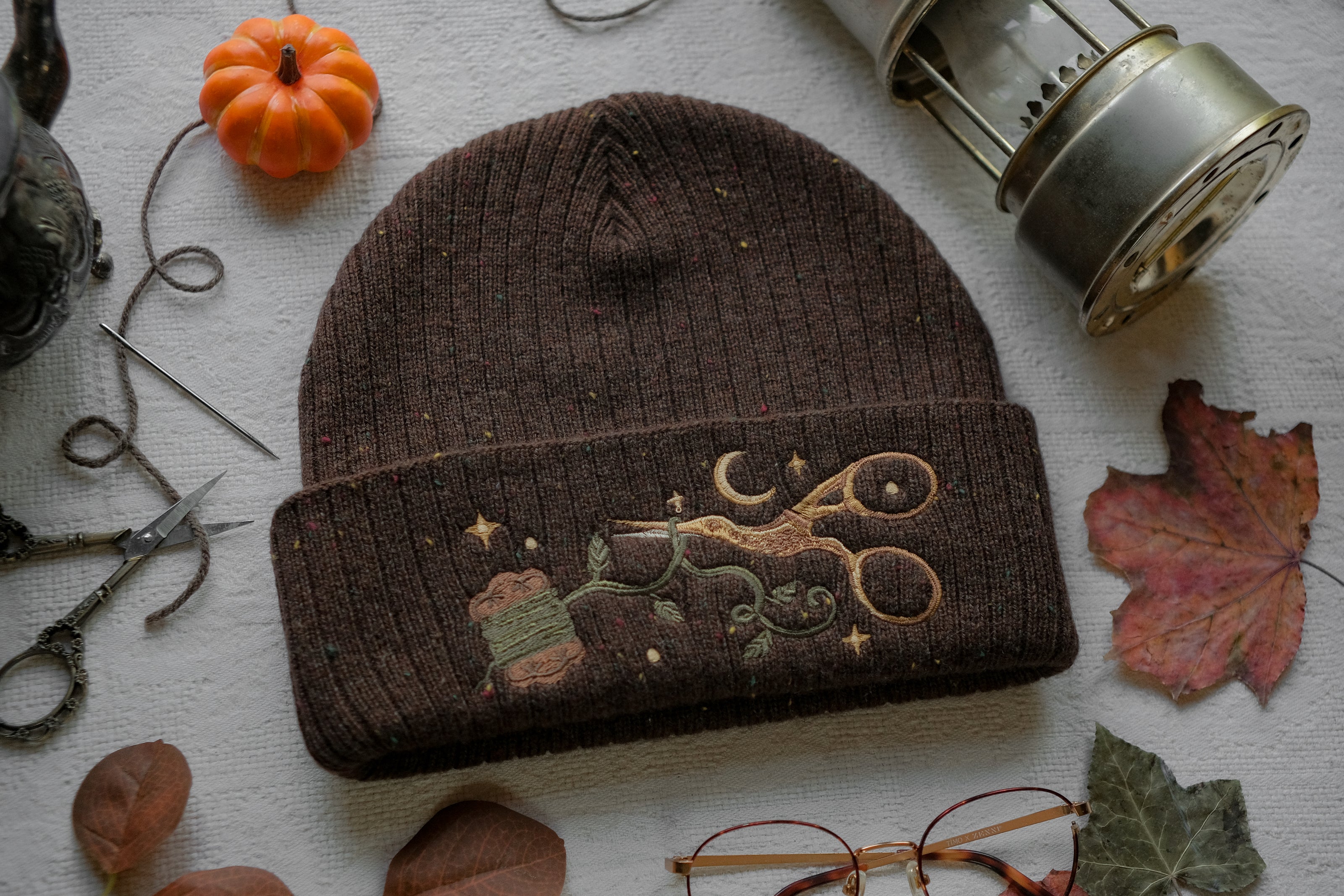 A cozy, flecked oak-brown, wool blend knit beanie, embroidered with a pair of golden, stork-shaped sewing scissors, and looping bobbin of green thread, under a crescent moon