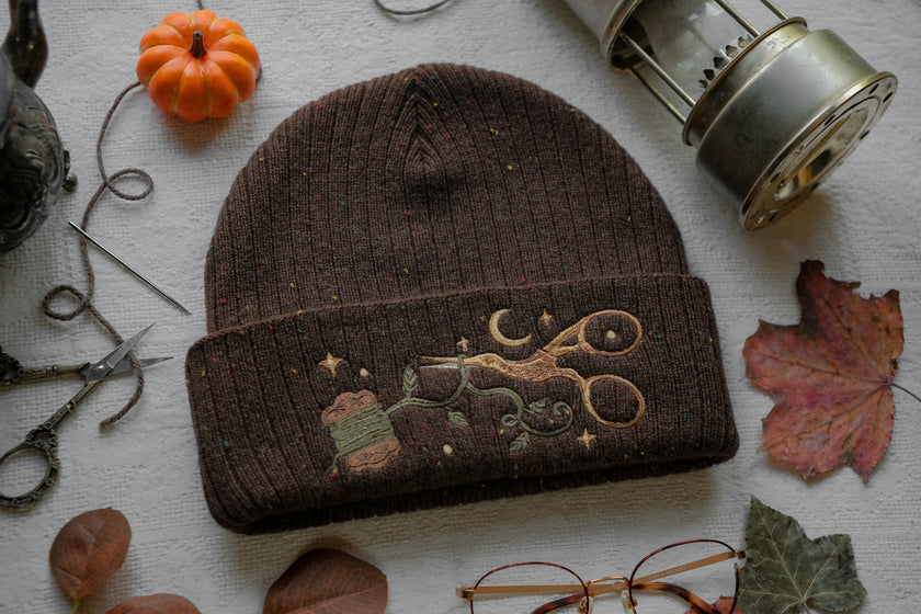 A cozy, flecked oak-brown, wool blend knit beanie, embroidered with a pair of golden, stork-shaped sewing scissors, and looping bobbin of green thread, under a crescent moon
