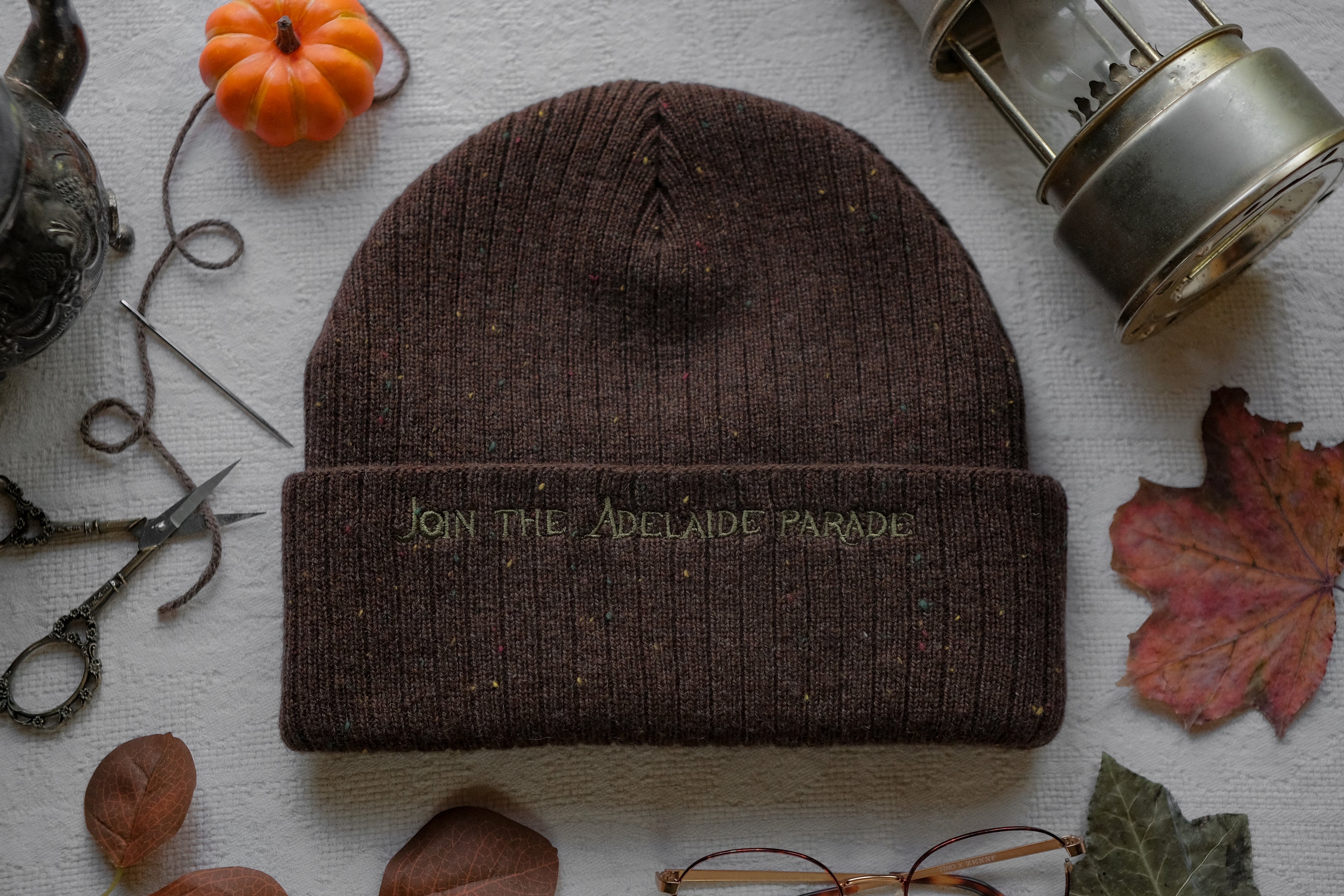 A cozy, flecked oak-brown, wool blend knit beanie, embroidered with a pair of golden, stork-shaped sewing scissors, and looping bobbin of green thread, under a crescent moon