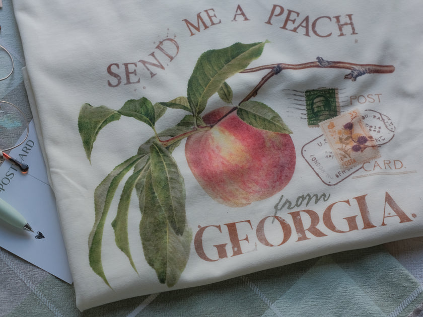 A cotton, vintage-style ringer t-shirt, in shades of soft cream and sage. The shirt is printed with a faded vintage peach design with postage stamps, and reads â€œsend me a peach from Georgiaâ€.