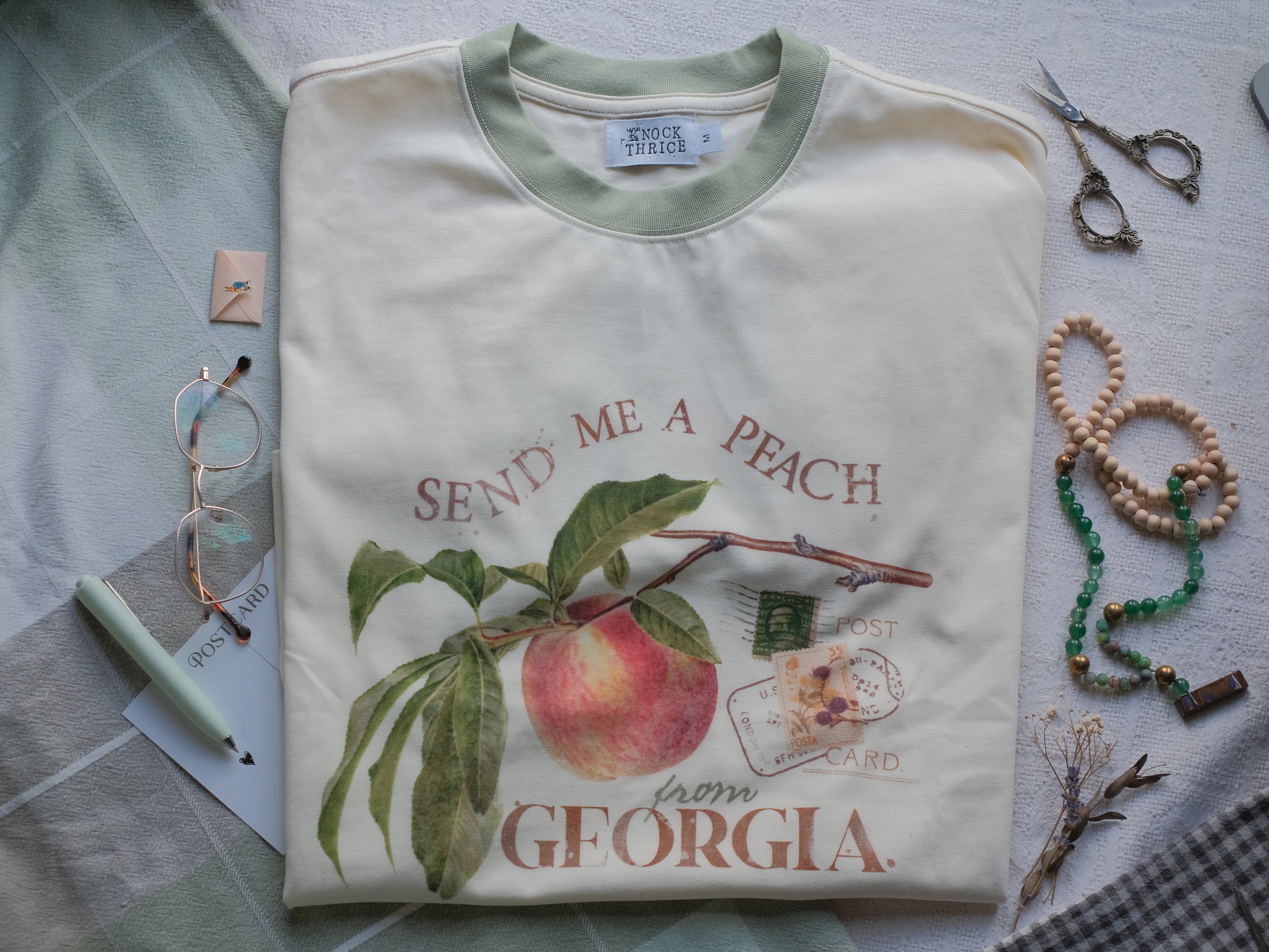 A cotton, vintage-style ringer t-shirt, in shades of soft cream and sage. The shirt is printed with a faded vintage peach design with postage stamps, and reads â€œsend me a peach from Georgiaâ€.