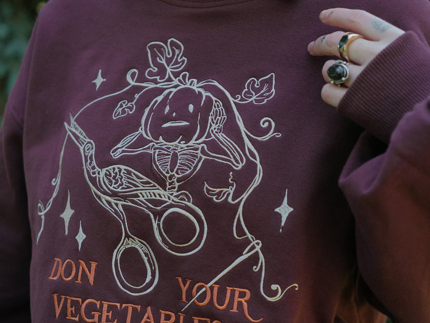 Cozy, oversized, puff-sleeved, cotton with fleece-lining, crewneck sweater in burgundy. The sweater features an intricately embroidered design in white and orange, of a pumpkin skeleton, vines, stork scissors, and reads â€œDon Your Vegetablesâ€.