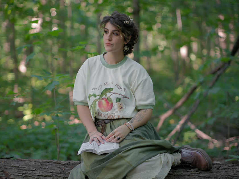 A cotton, vintage-style ringer t-shirt, in shades of soft cream and sage. The shirt is printed with a faded vintage peach design with postage stamps, and reads â€œsend me a peach from Georgiaâ€.