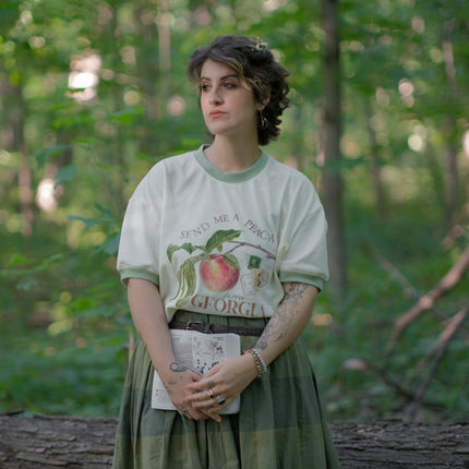 A cotton, vintage-style ringer t-shirt, in shades of soft cream and sage. The shirt is printed with a faded vintage peach design with postage stamps, and reads â€œsend me a peach from Georgiaâ€.
