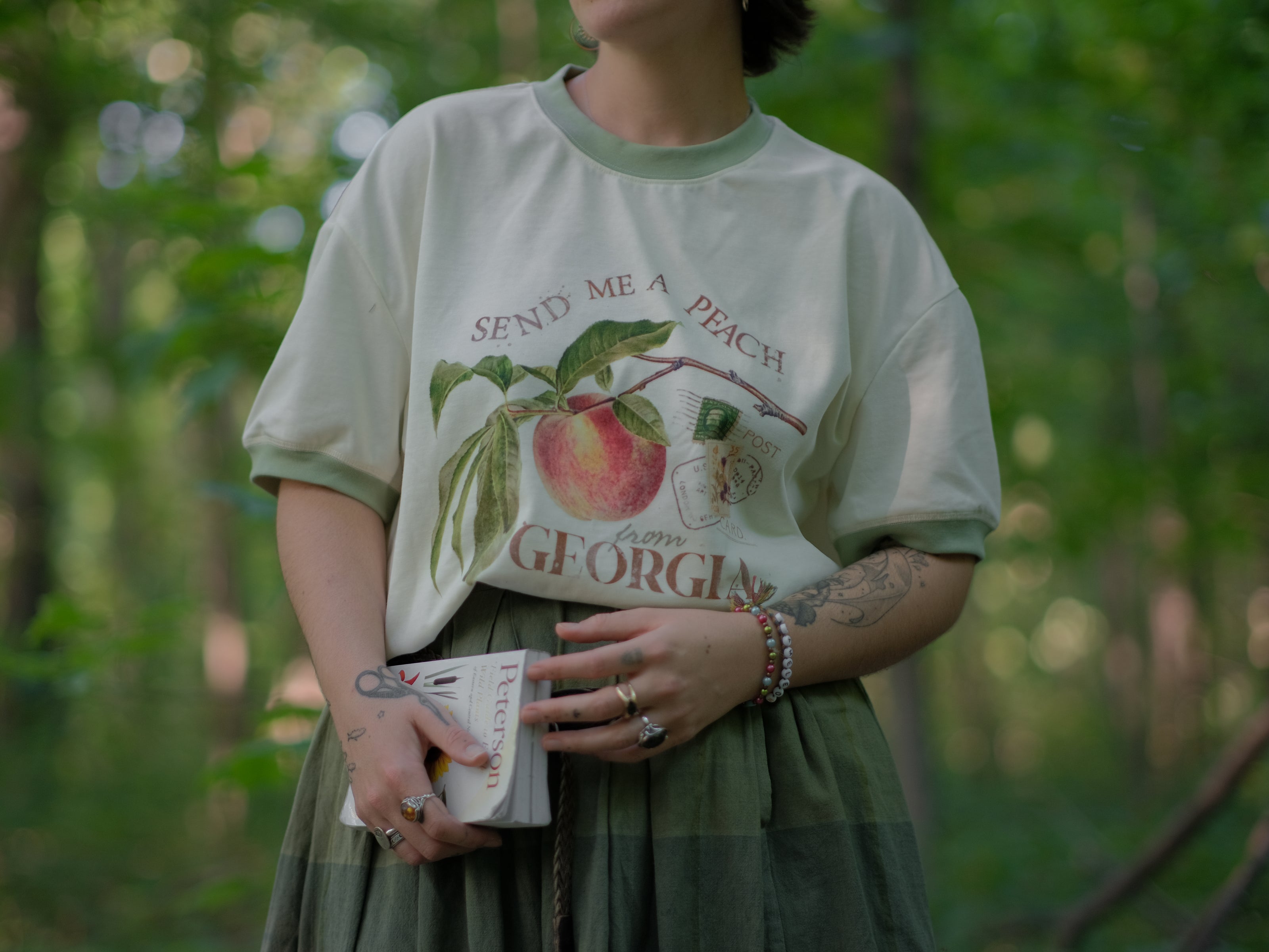 A cotton, vintage-style ringer t-shirt, in shades of soft cream and sage. The shirt is printed with a faded vintage peach design with postage stamps, and reads â€œsend me a peach from Georgiaâ€.
