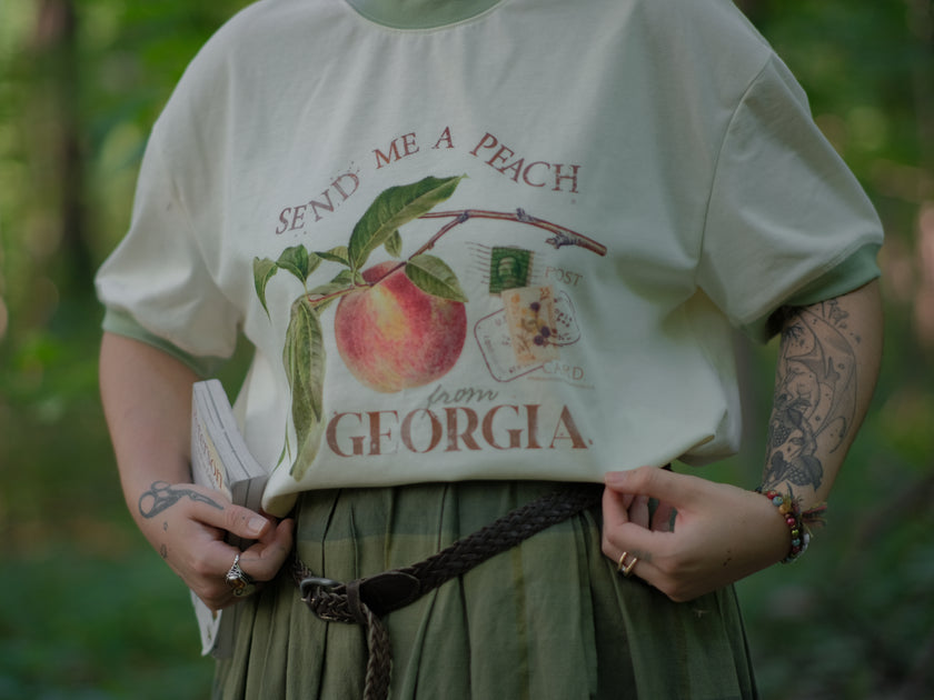 A cotton, vintage-style ringer t-shirt, in shades of soft cream and sage. The shirt is printed with a faded vintage peach design with postage stamps, and reads â€œsend me a peach from Georgiaâ€.