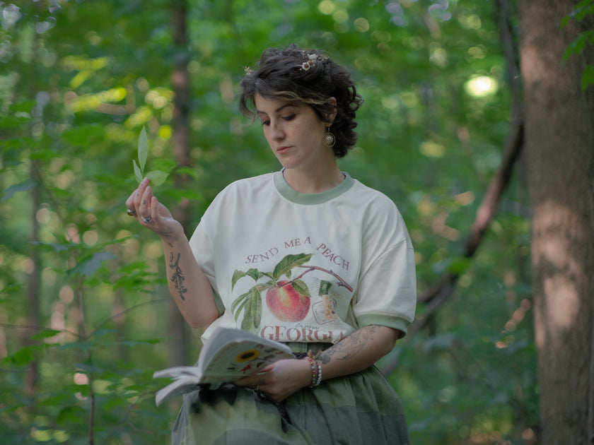 A cotton, vintage-style ringer t-shirt, in shades of soft cream and sage. The shirt is printed with a faded vintage peach design with postage stamps, and reads â€œsend me a peach from Georgiaâ€.