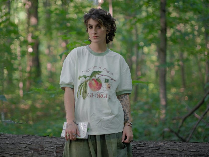 A cotton, vintage-style ringer t-shirt, in shades of soft cream and sage. The shirt is printed with a faded vintage peach design with postage stamps, and reads â€œsend me a peach from Georgiaâ€.