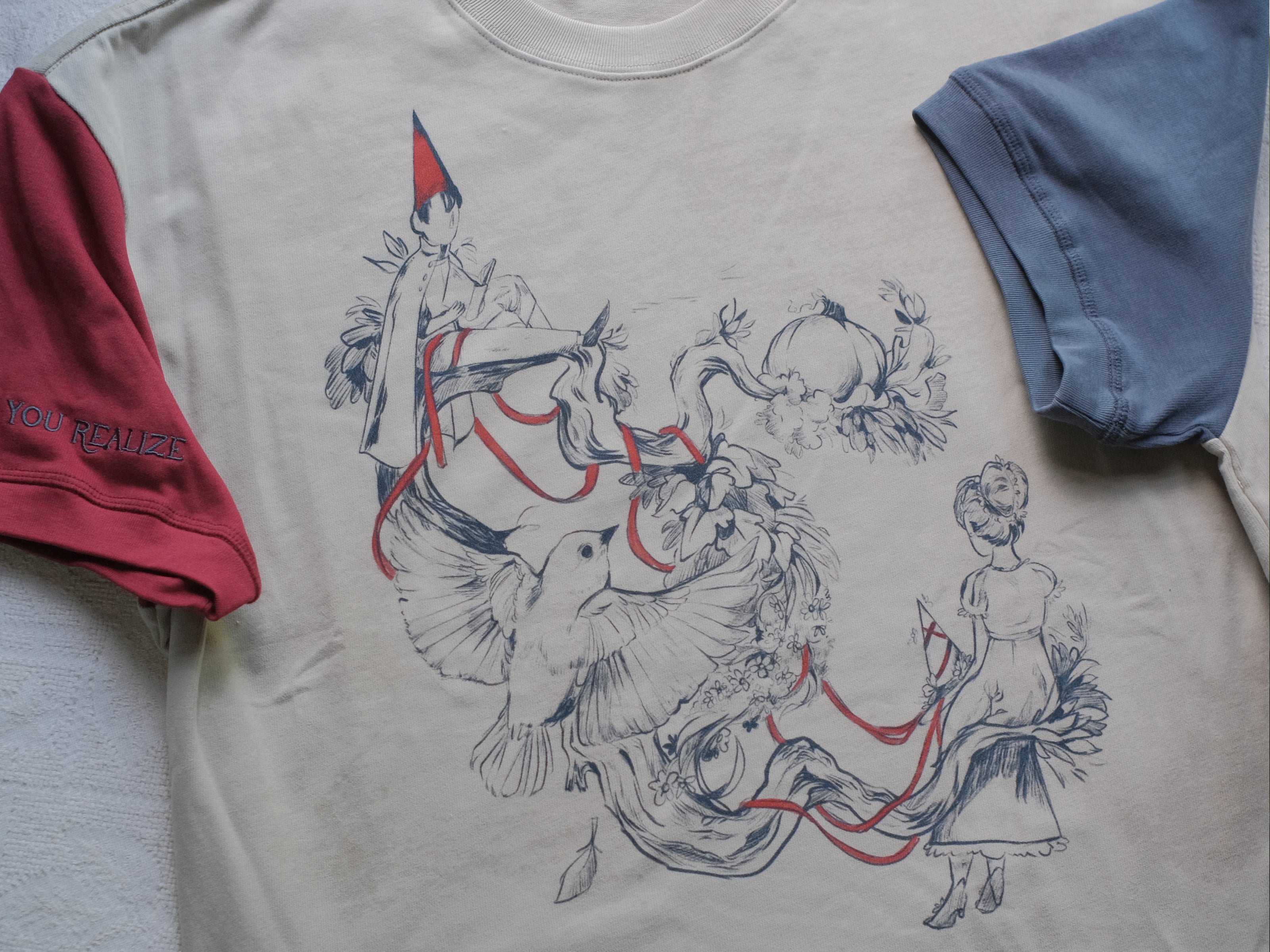 A cotton, vintage-style ringer t-shirt, in soft cream, and mismatched sleeves in red and navy. The shirt is printed with a tapestry inspired illustration including details of some young-lovers, a ripe pumpkin, sturdy tree branches, and a red ribbon.