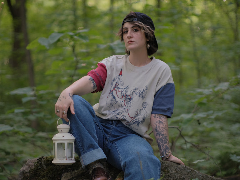 A cotton, vintage-style ringer t-shirt, in soft cream, and mismatched sleeves in red and navy. The shirt is printed with a tapestry inspired illustration including details of some young-lovers, a ripe pumpkin, sturdy tree branches, and a red ribbon.