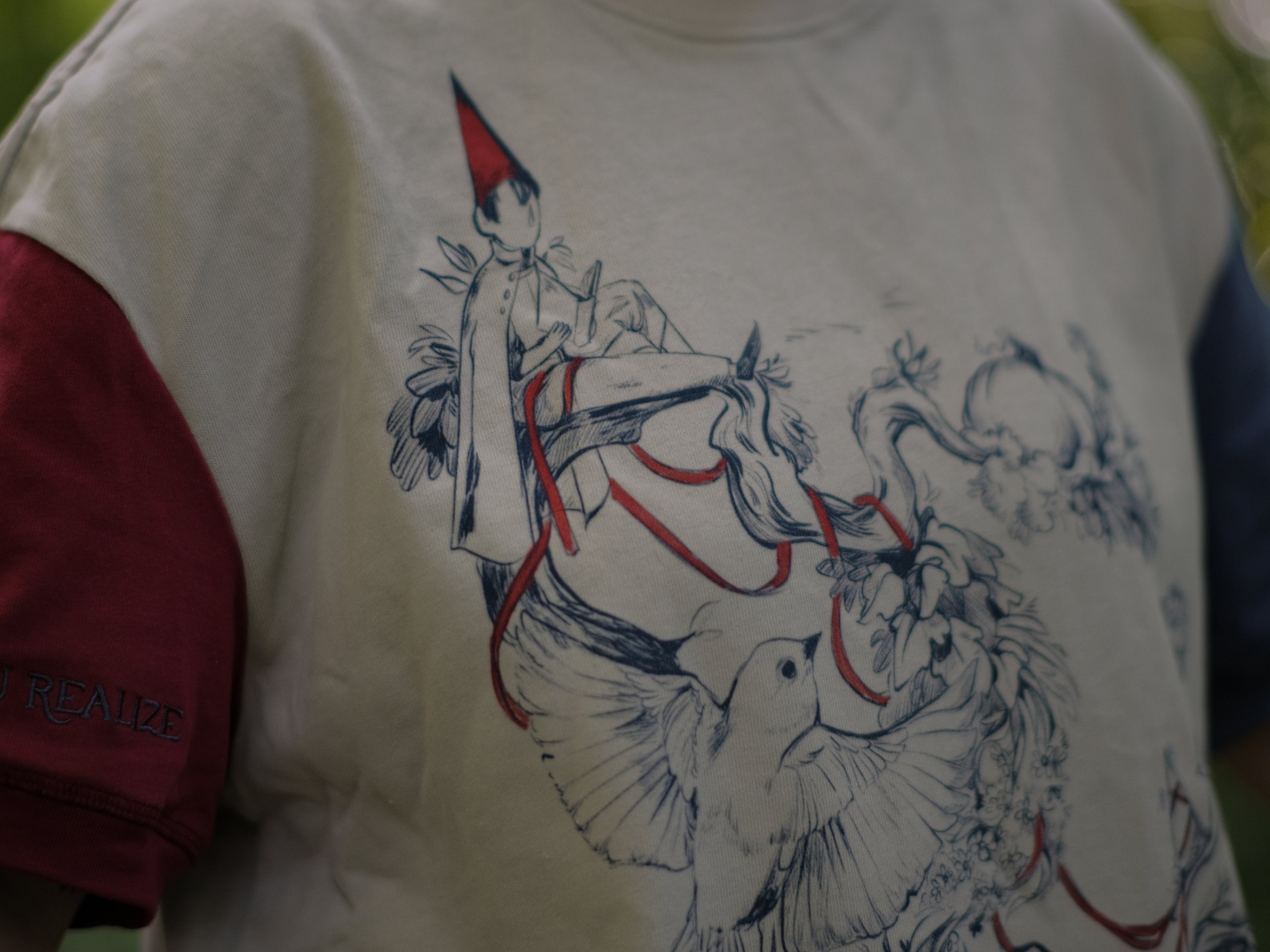 A cotton, vintage-style ringer t-shirt, in soft cream, and mismatched sleeves in red and navy. The shirt is printed with a tapestry inspired illustration including details of some young-lovers, a ripe pumpkin, sturdy tree branches, and a red ribbon.
