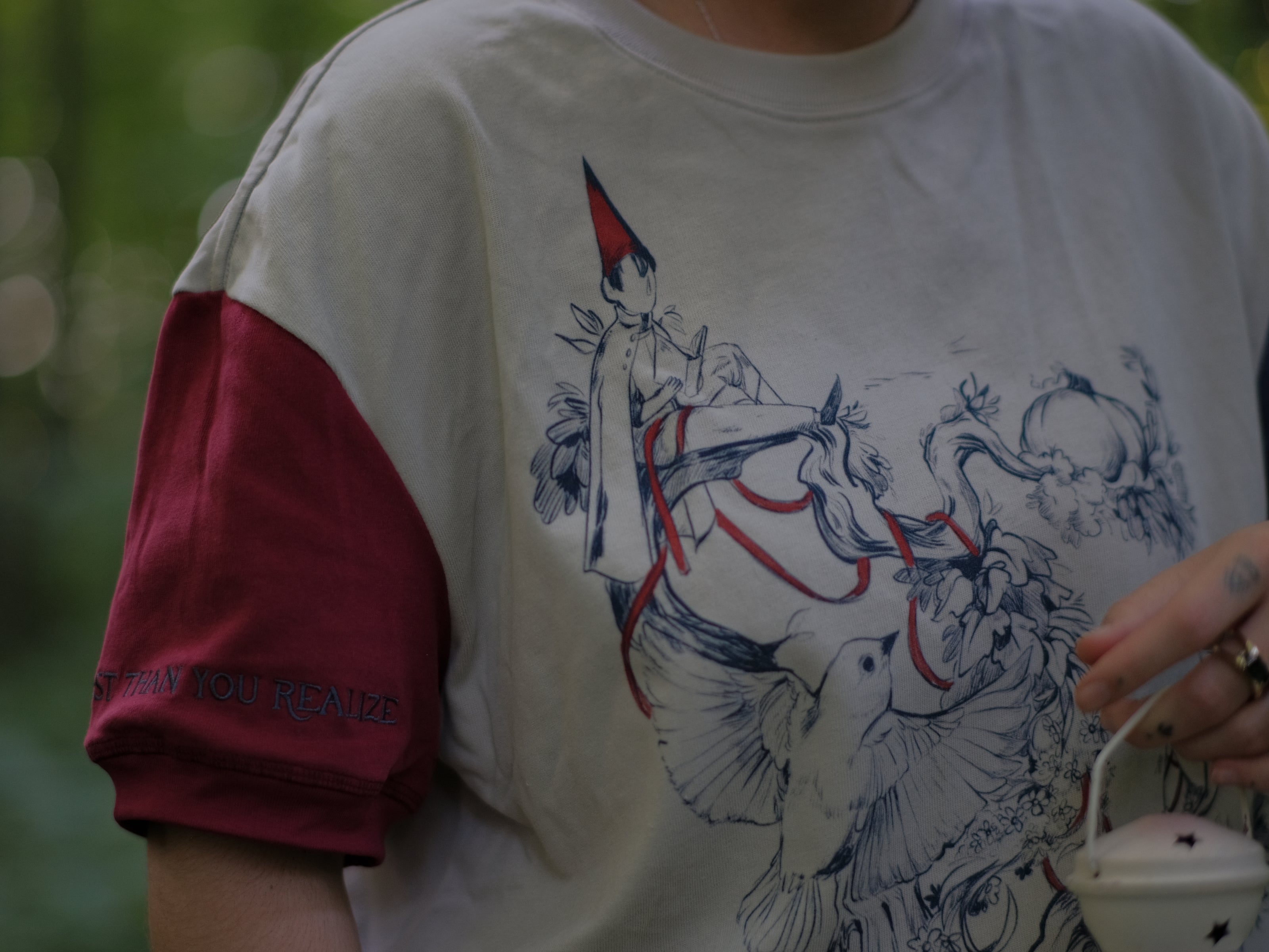 A cotton, vintage-style ringer t-shirt, in soft cream, and mismatched sleeves in red and navy. The shirt is printed with a tapestry inspired illustration including details of some young-lovers, a ripe pumpkin, sturdy tree branches, and a red ribbon.