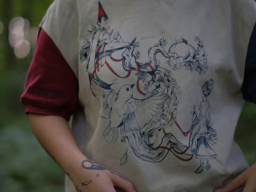 A cotton, vintage-style ringer t-shirt, in soft cream, and mismatched sleeves in red and navy. The shirt is printed with a tapestry inspired illustration including details of some young-lovers, a ripe pumpkin, sturdy tree branches, and a red ribbon.