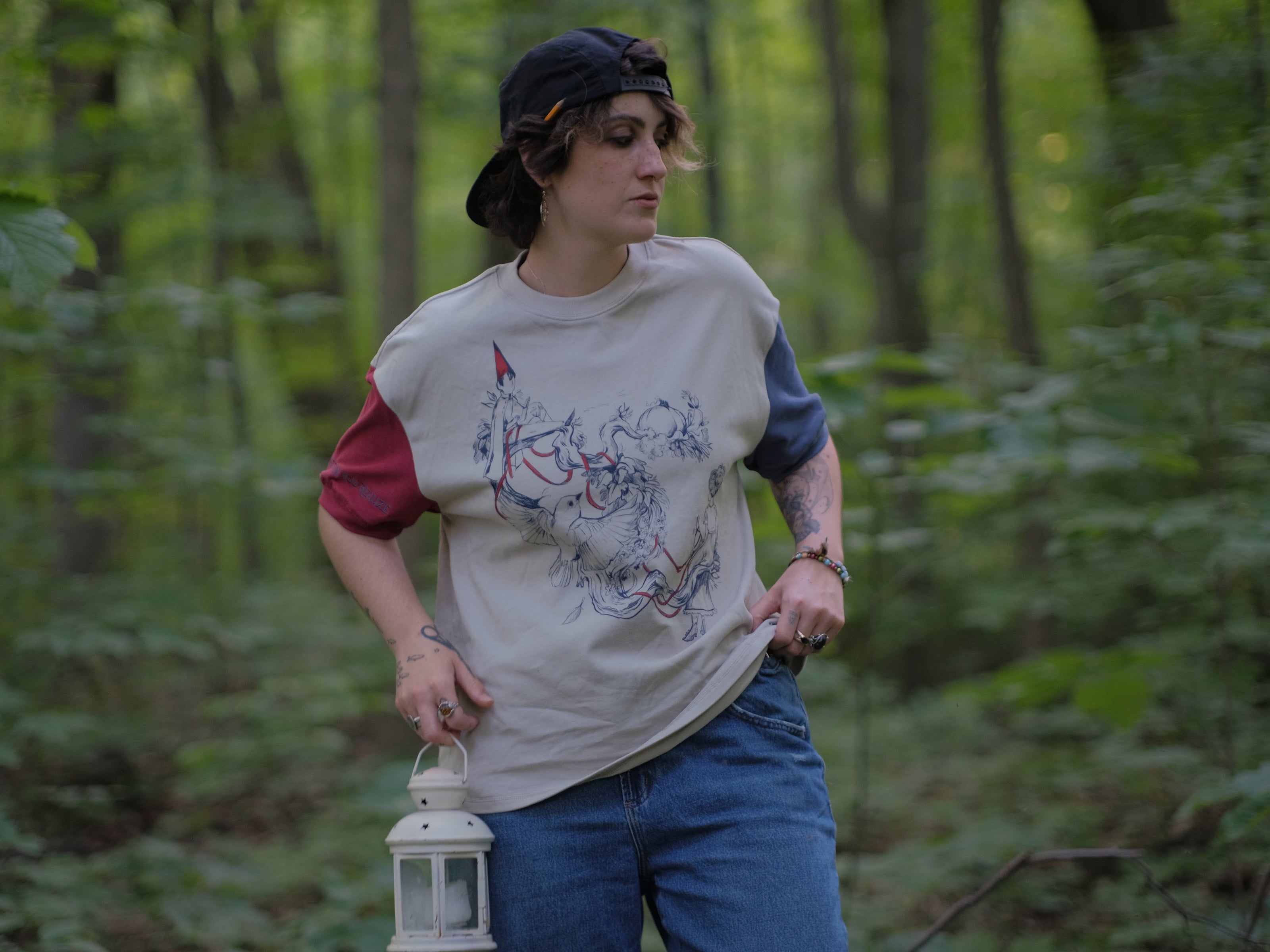 A cotton, vintage-style ringer t-shirt, in soft cream, and mismatched sleeves in red and navy. The shirt is printed with a tapestry inspired illustration including details of some young-lovers, a ripe pumpkin, sturdy tree branches, and a red ribbon.