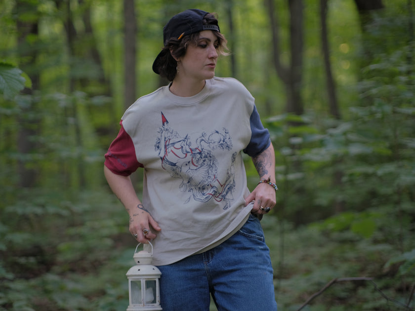 A cotton, vintage-style ringer t-shirt, in soft cream, and mismatched sleeves in red and navy. The shirt is printed with a tapestry inspired illustration including details of some young-lovers, a ripe pumpkin, sturdy tree branches, and a red ribbon.