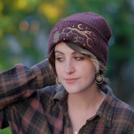 A cozy, flecked, amaranth-red, wool blend knit beanie, embroidered with a pair of golden, stork-shaped sewing scissors, and looping bobbin of green thread, under a crescent moon.