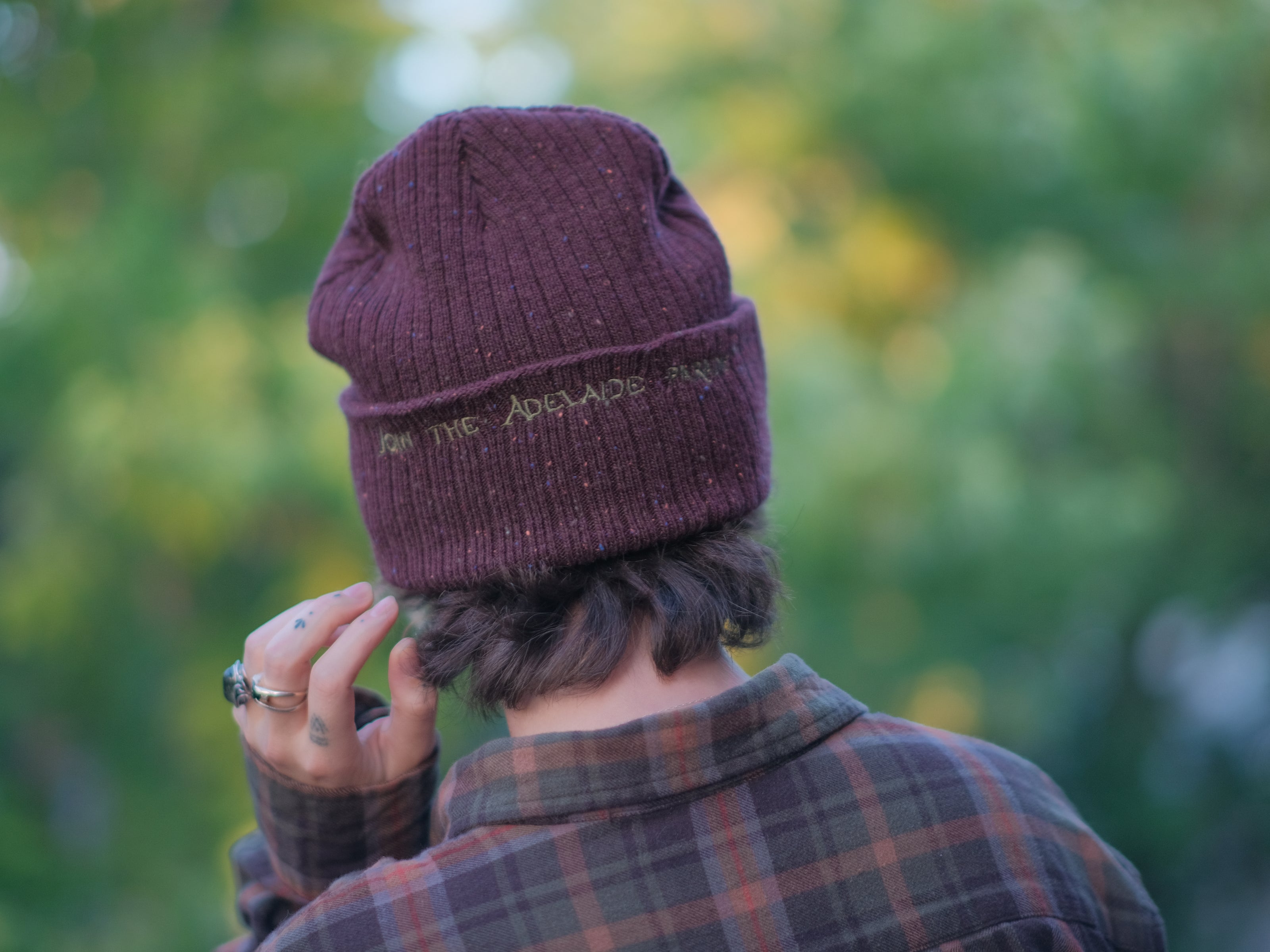 A cozy, flecked, amaranth-red, wool blend knit beanie, embroidered with a pair of golden, stork-shaped sewing scissors, and looping bobbin of green thread, under a crescent moon.