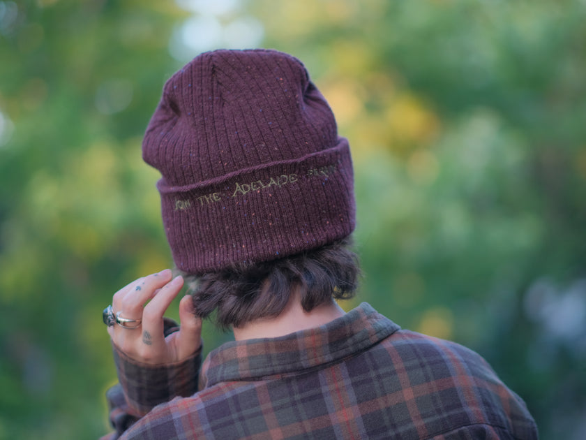 A cozy, flecked, amaranth-red, wool blend knit beanie, embroidered with a pair of golden, stork-shaped sewing scissors, and looping bobbin of green thread, under a crescent moon.