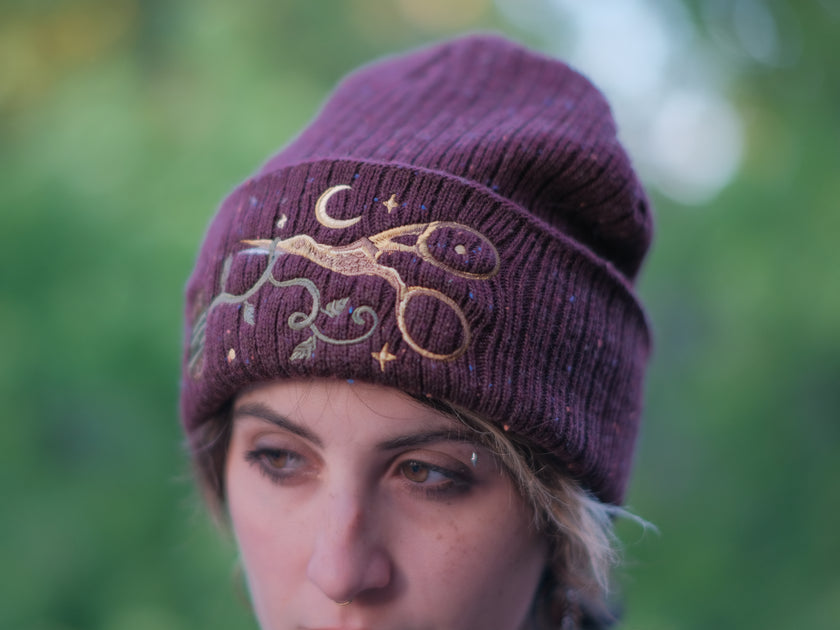 A cozy, flecked, amaranth-red, wool blend knit beanie, embroidered with a pair of golden, stork-shaped sewing scissors, and looping bobbin of green thread, under a crescent moon.