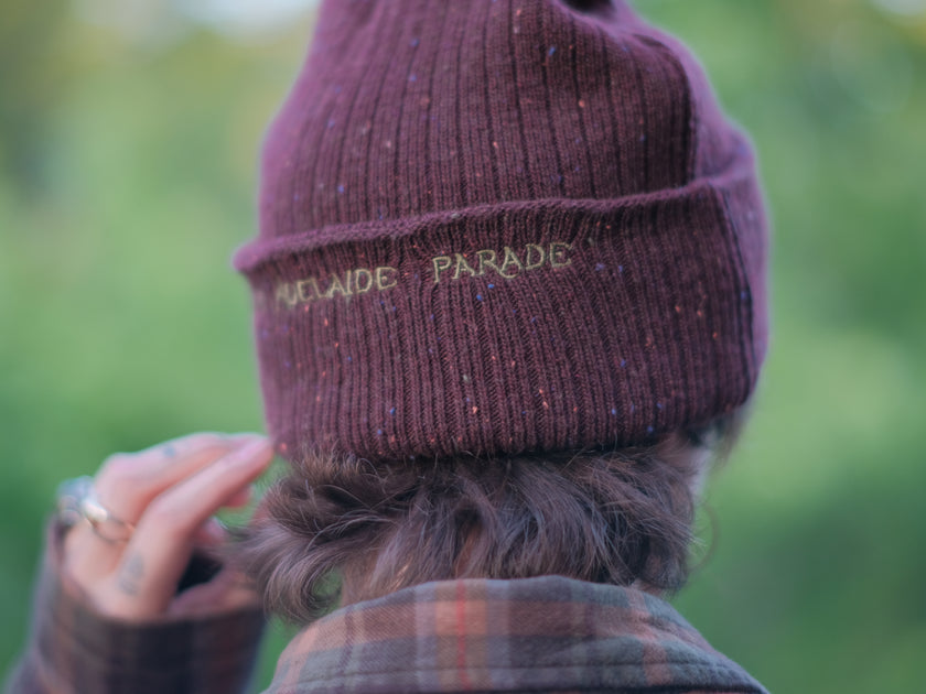 A cozy, flecked, amaranth-red, wool blend knit beanie, embroidered with a pair of golden, stork-shaped sewing scissors, and looping bobbin of green thread, under a crescent moon.