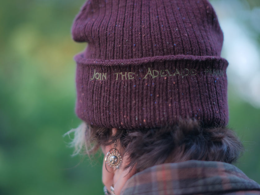 A cozy, flecked, amaranth-red, wool blend knit beanie, embroidered with a pair of golden, stork-shaped sewing scissors, and looping bobbin of green thread, under a crescent moon.