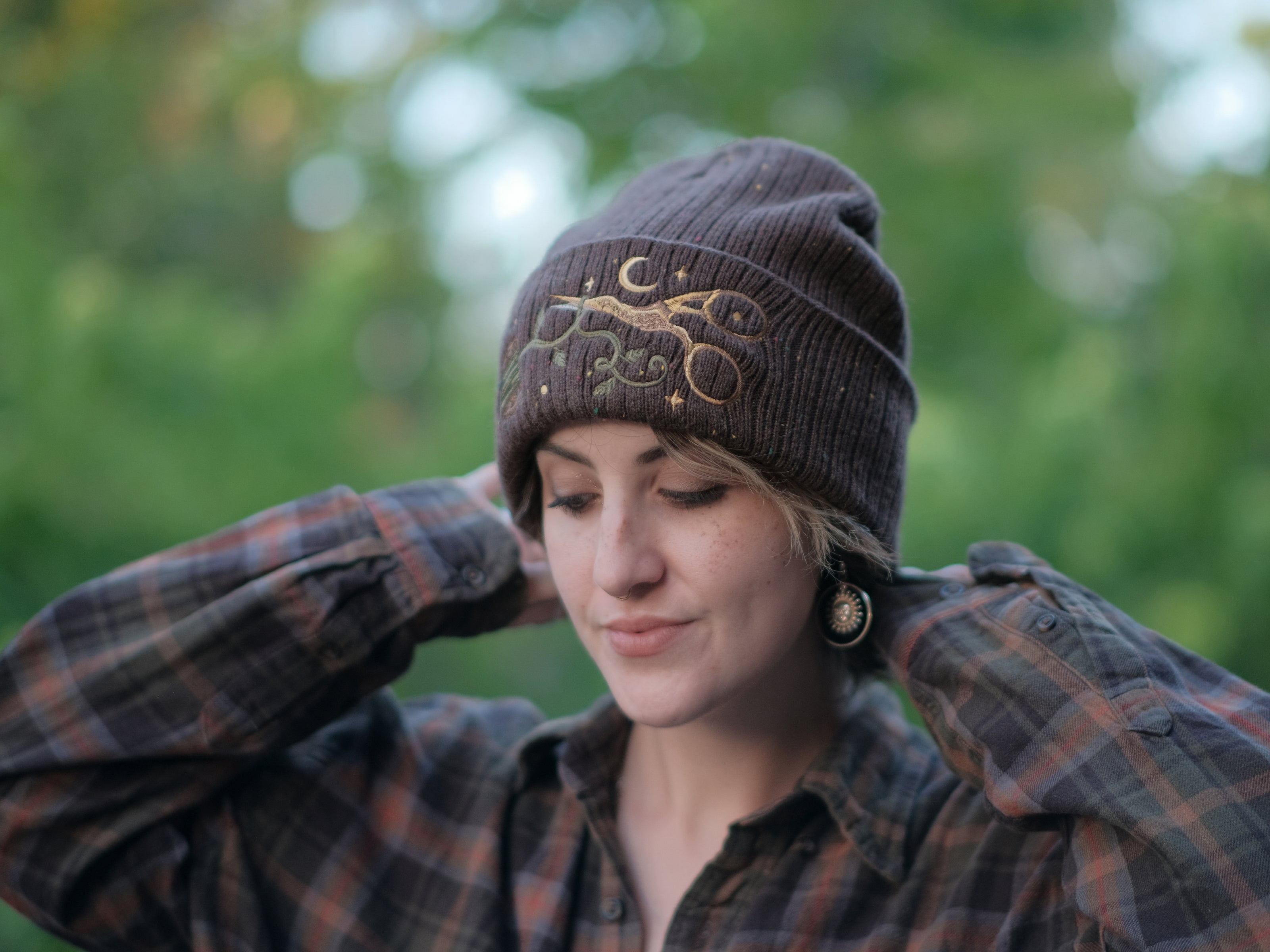 A cozy, flecked oak-brown, wool blend knit beanie, embroidered with a pair of golden, stork-shaped sewing scissors, and looping bobbin of green thread, under a crescent moon