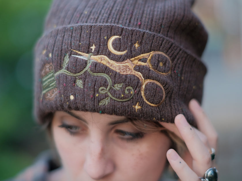 A cozy, flecked oak-brown, wool blend knit beanie, embroidered with a pair of golden, stork-shaped sewing scissors, and looping bobbin of green thread, under a crescent moon