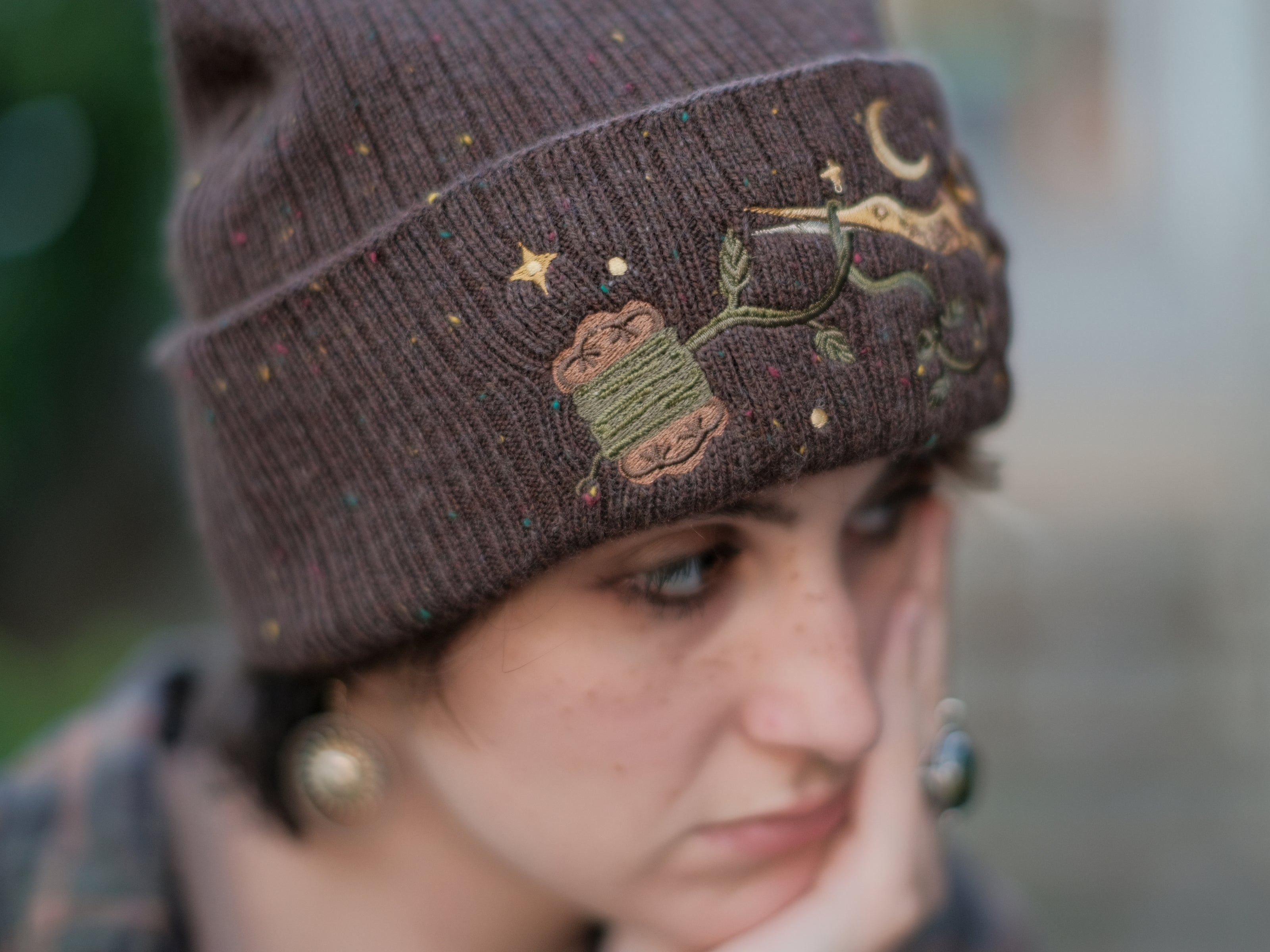 A cozy, flecked oak-brown, wool blend knit beanie, embroidered with a pair of golden, stork-shaped sewing scissors, and looping bobbin of green thread, under a crescent moon