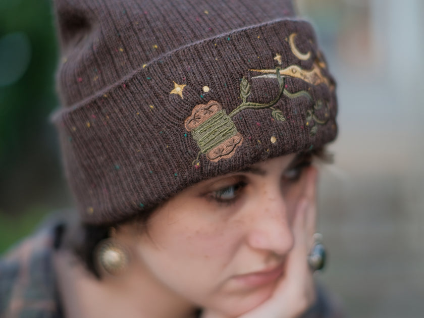 A cozy, flecked oak-brown, wool blend knit beanie, embroidered with a pair of golden, stork-shaped sewing scissors, and looping bobbin of green thread, under a crescent moon