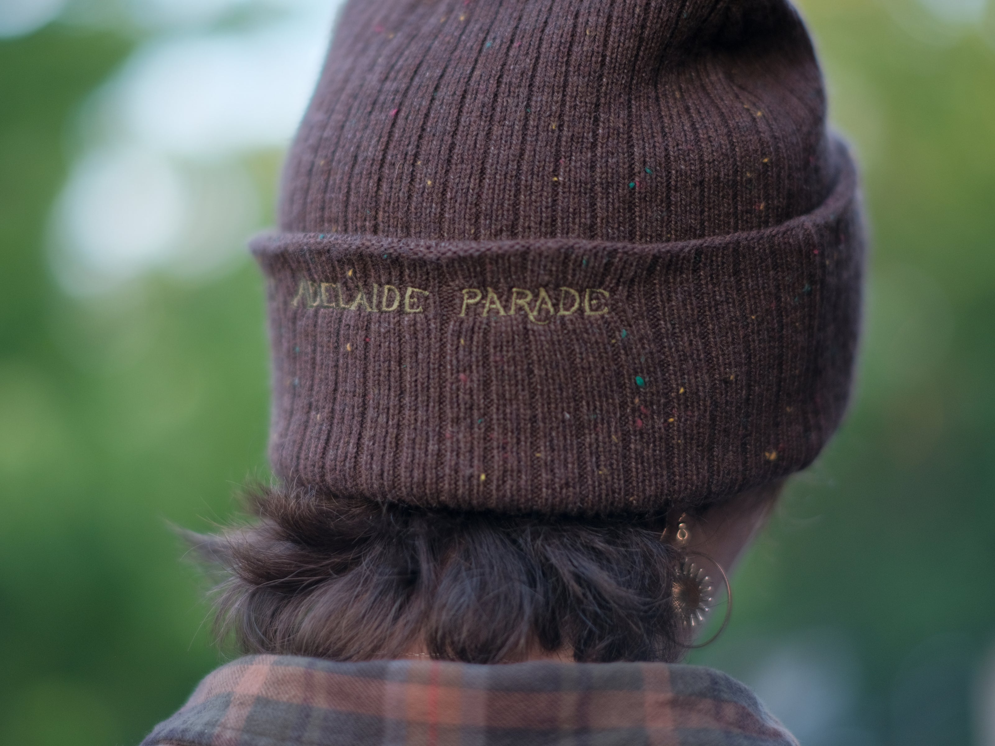 A cozy, flecked oak-brown, wool blend knit beanie, embroidered with a pair of golden, stork-shaped sewing scissors, and looping bobbin of green thread, under a crescent moon