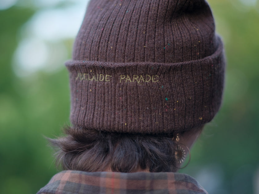 A cozy, flecked oak-brown, wool blend knit beanie, embroidered with a pair of golden, stork-shaped sewing scissors, and looping bobbin of green thread, under a crescent moon