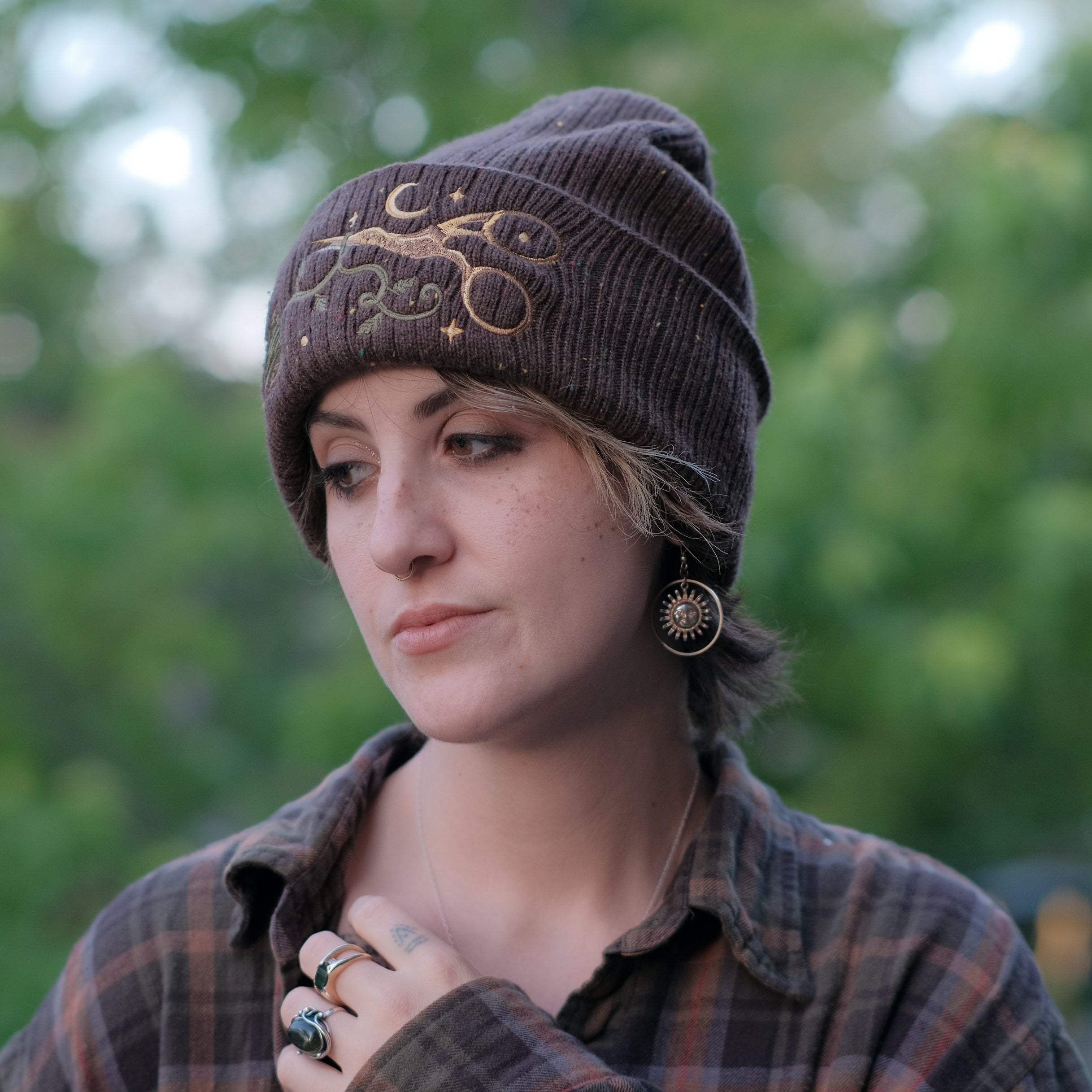 A cozy, flecked oak-brown, wool blend knit beanie, embroidered with a pair of golden, stork-shaped sewing scissors, and looping bobbin of green thread, under a crescent moon