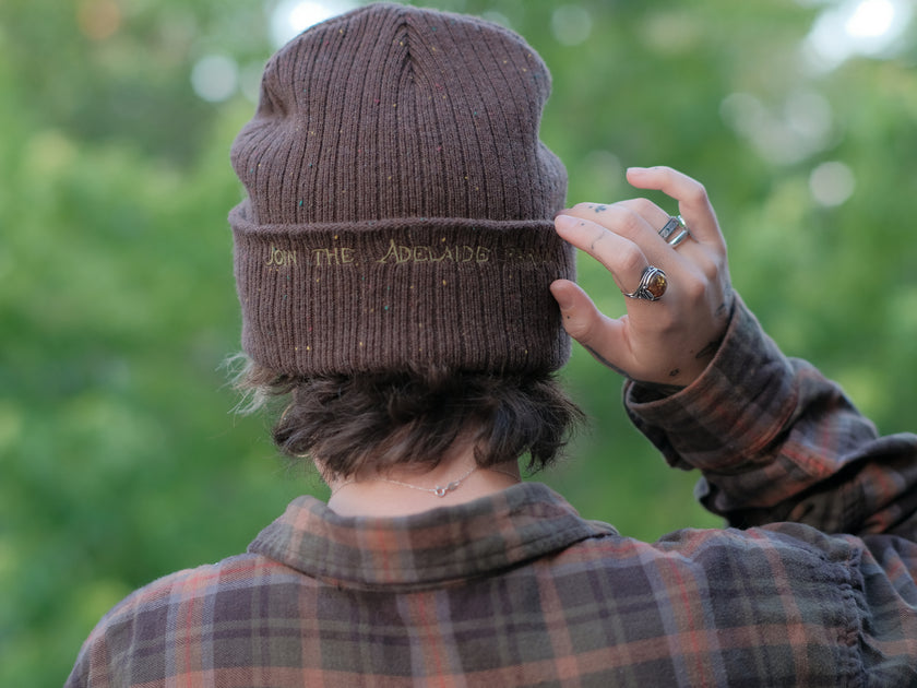 A cozy, flecked oak-brown, wool blend knit beanie, embroidered with a pair of golden, stork-shaped sewing scissors, and looping bobbin of green thread, under a crescent moon
