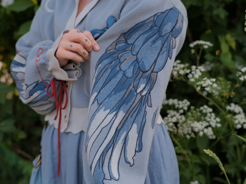A soft, knit, cropped cardigan, in tones of dove-grey and dusty-blue. This cardigan features detailed embroidery of a bow, moon and stars, stork scissors, and bluebird wings on both sleeves. The cardigan also features red-ribbon drawstring sleeves for customization, and scalloped edge detailing.