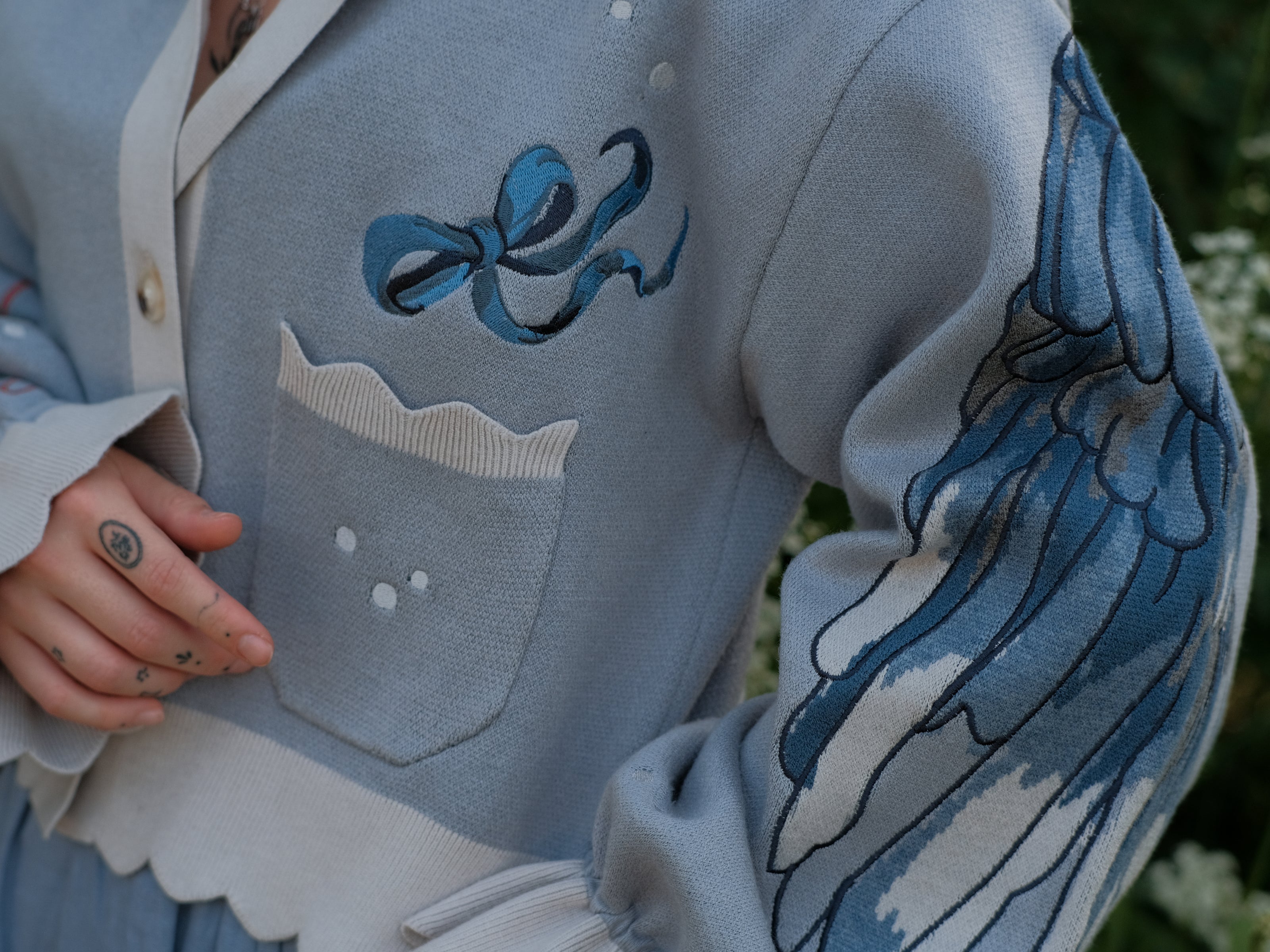 A soft, knit, cropped cardigan, in tones of dove-grey and dusty-blue. This cardigan features detailed embroidery of a bow, moon and stars, stork scissors, and bluebird wings on both sleeves. The cardigan also features red-ribbon drawstring sleeves for customization, and scalloped edge detailing.