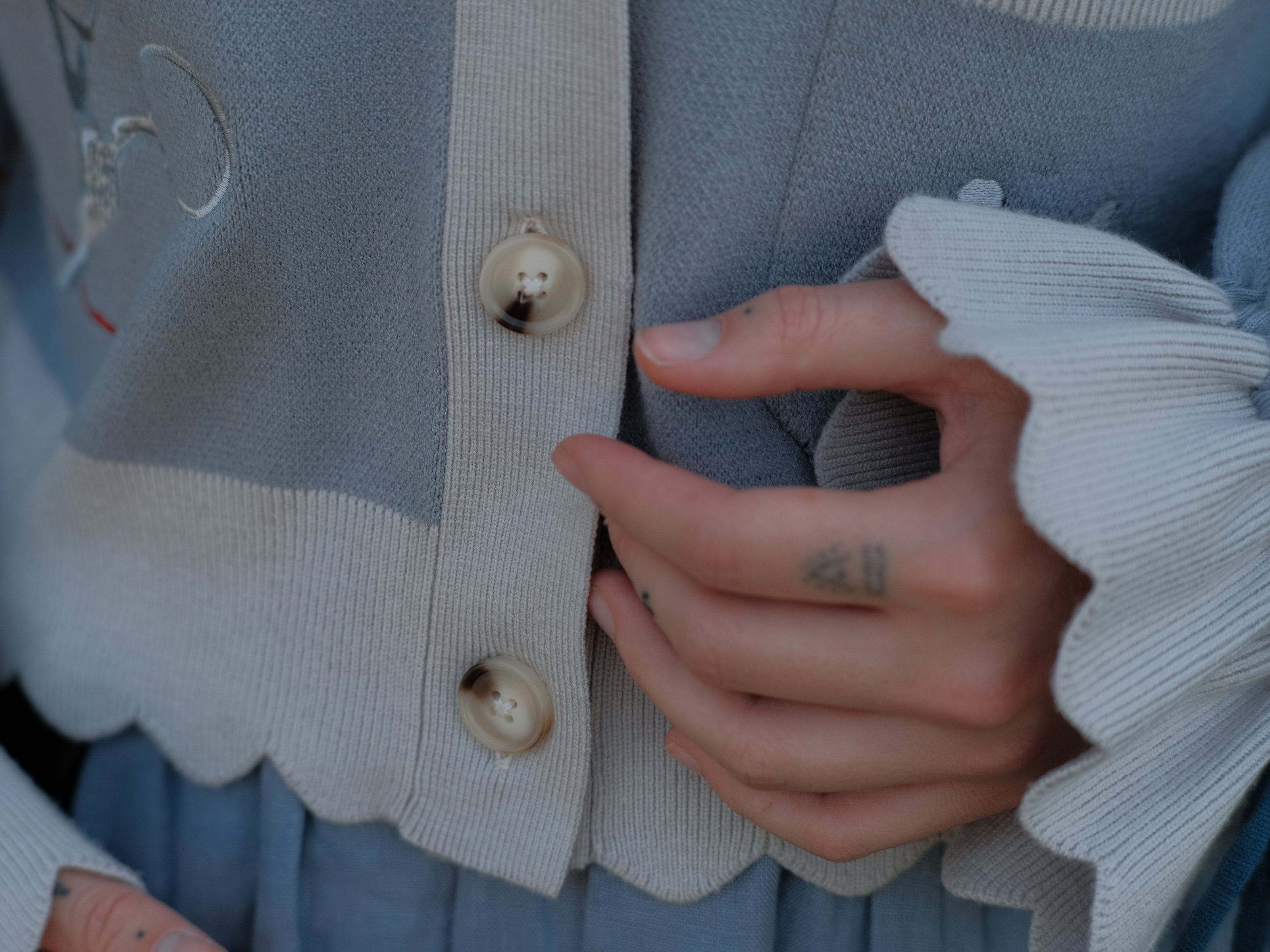 A soft, knit, cropped cardigan, in tones of dove-grey and dusty-blue. This cardigan features detailed embroidery of a bow, moon and stars, stork scissors, and bluebird wings on both sleeves. The cardigan also features red-ribbon drawstring sleeves for customization, and scalloped edge detailing.