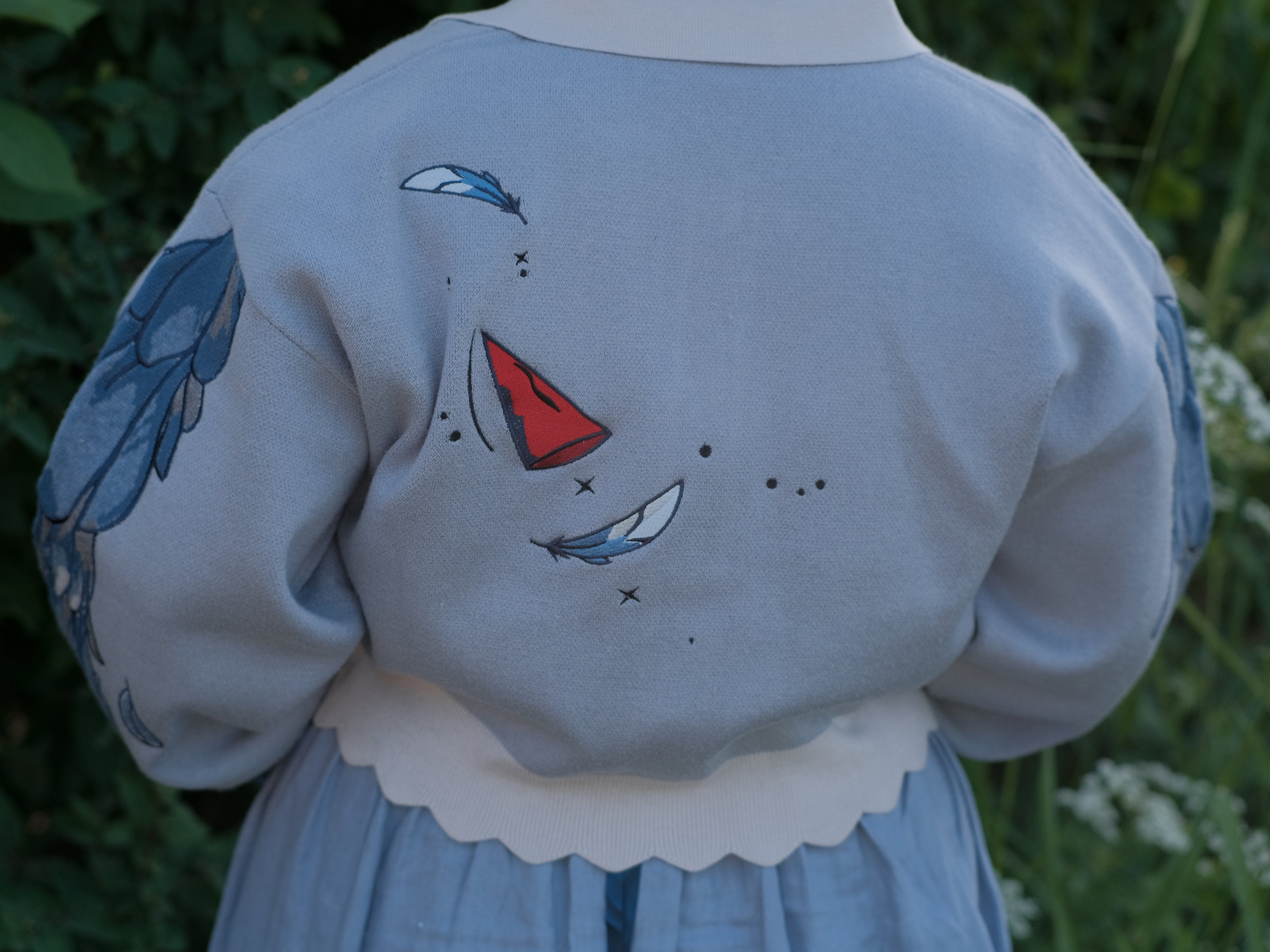 A soft, knit, cropped cardigan, in tones of dove-grey and dusty-blue. This cardigan features detailed embroidery of a bow, moon and stars, stork scissors, and bluebird wings on both sleeves. The cardigan also features red-ribbon drawstring sleeves for customization, and scalloped edge detailing.