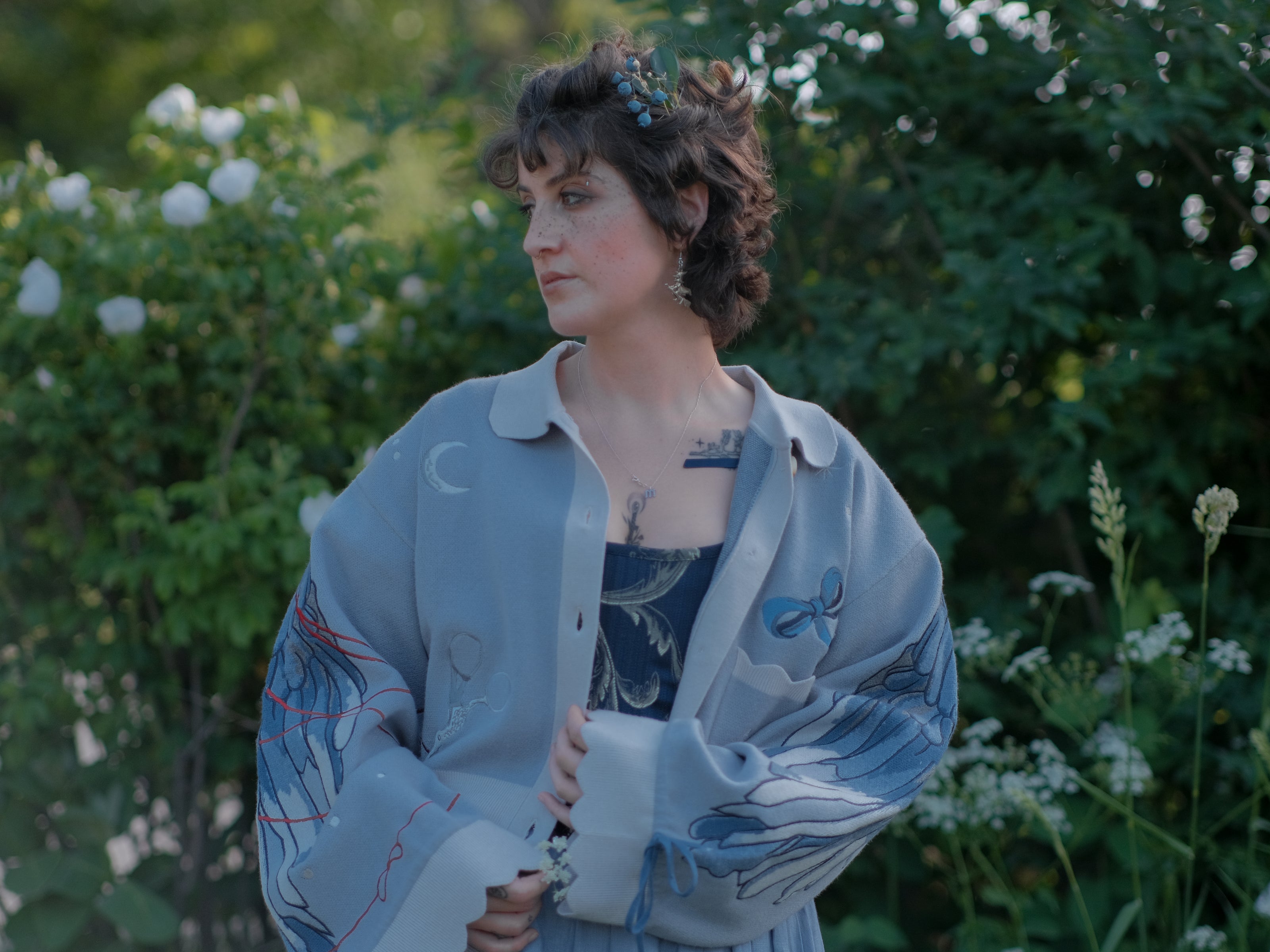 A soft, knit, cropped cardigan, in tones of dove-grey and dusty-blue. This cardigan features detailed embroidery of a bow, moon and stars, stork scissors, and bluebird wings on both sleeves. The cardigan also features red-ribbon drawstring sleeves for customization, and scalloped edge detailing.