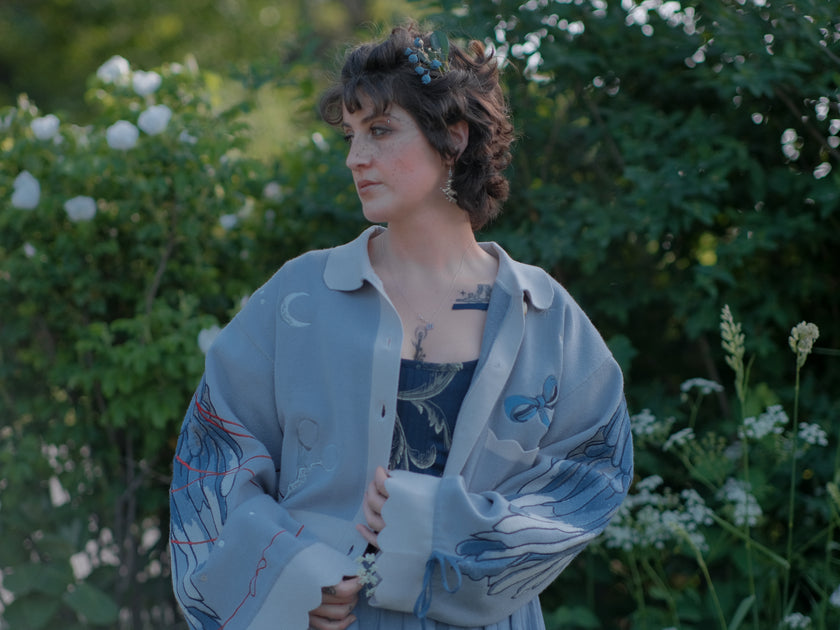A soft, knit, cropped cardigan, in tones of dove-grey and dusty-blue. This cardigan features detailed embroidery of a bow, moon and stars, stork scissors, and bluebird wings on both sleeves. The cardigan also features red-ribbon drawstring sleeves for customization, and scalloped edge detailing.
