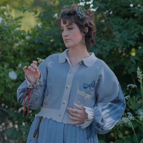 A soft, knit, cropped cardigan, in tones of dove-grey and dusty-blue. This cardigan features detailed embroidery of a bow, moon and stars, stork scissors, and bluebird wings on both sleeves. The cardigan also features red-ribbon drawstring sleeves for customization, and scalloped edge detailing.