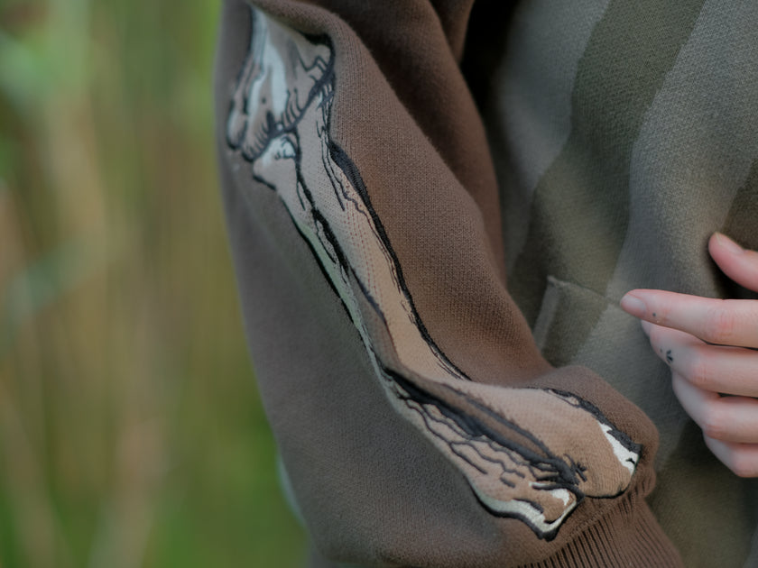 Cozy, cotton, knit hoodie featuring a striped body in shades of green, brown skeleton embroidered sleeves, and an orange pumpkin hood. The front features an embroidered harvest badge, and the back reads â€œyouâ€™ll join us somedayâ€.