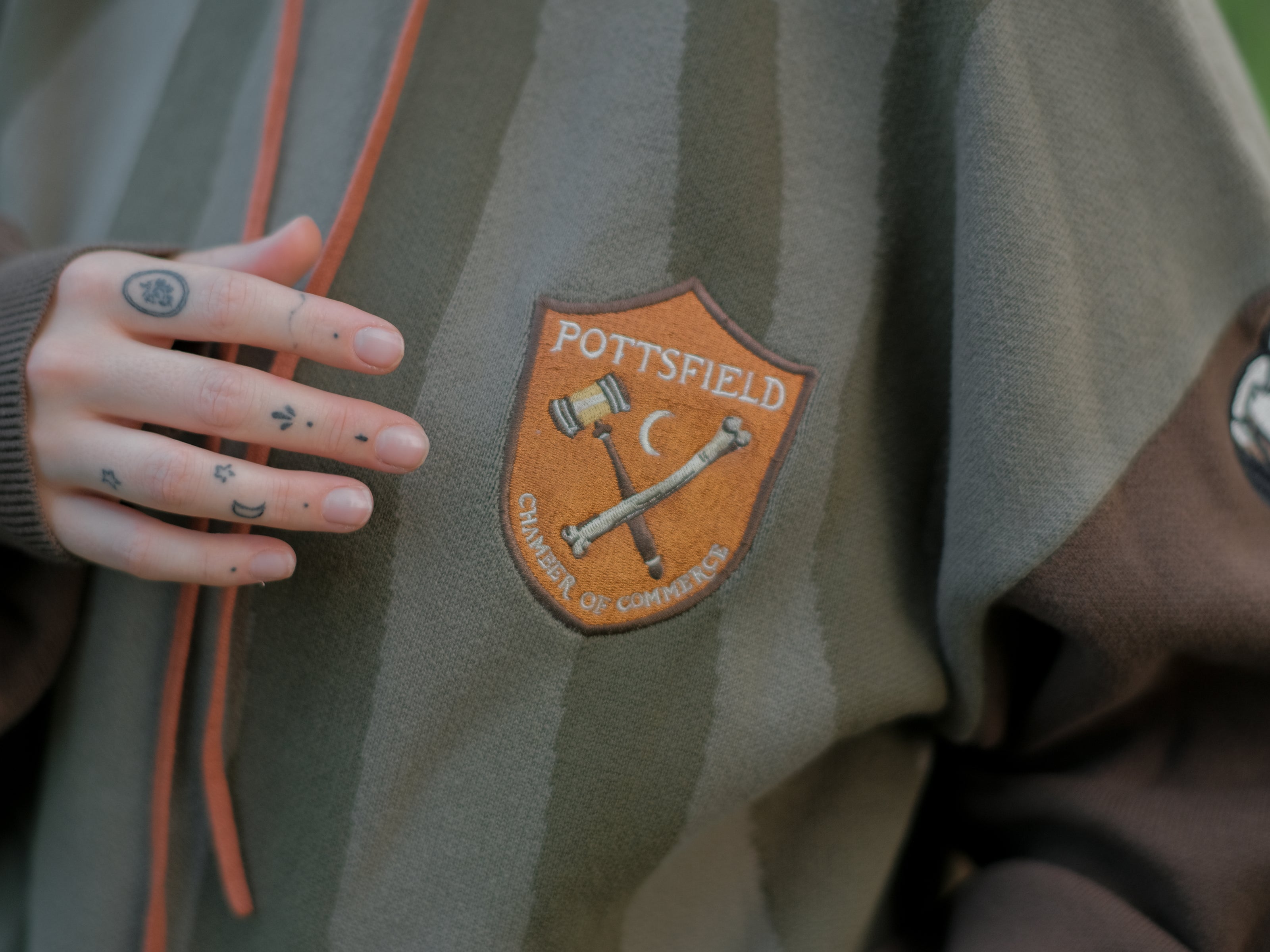 Cozy, cotton, knit hoodie featuring a striped body in shades of green, brown skeleton embroidered sleeves, and an orange pumpkin hood. The front features an embroidered harvest badge, and the back reads â€œyouâ€™ll join us somedayâ€.